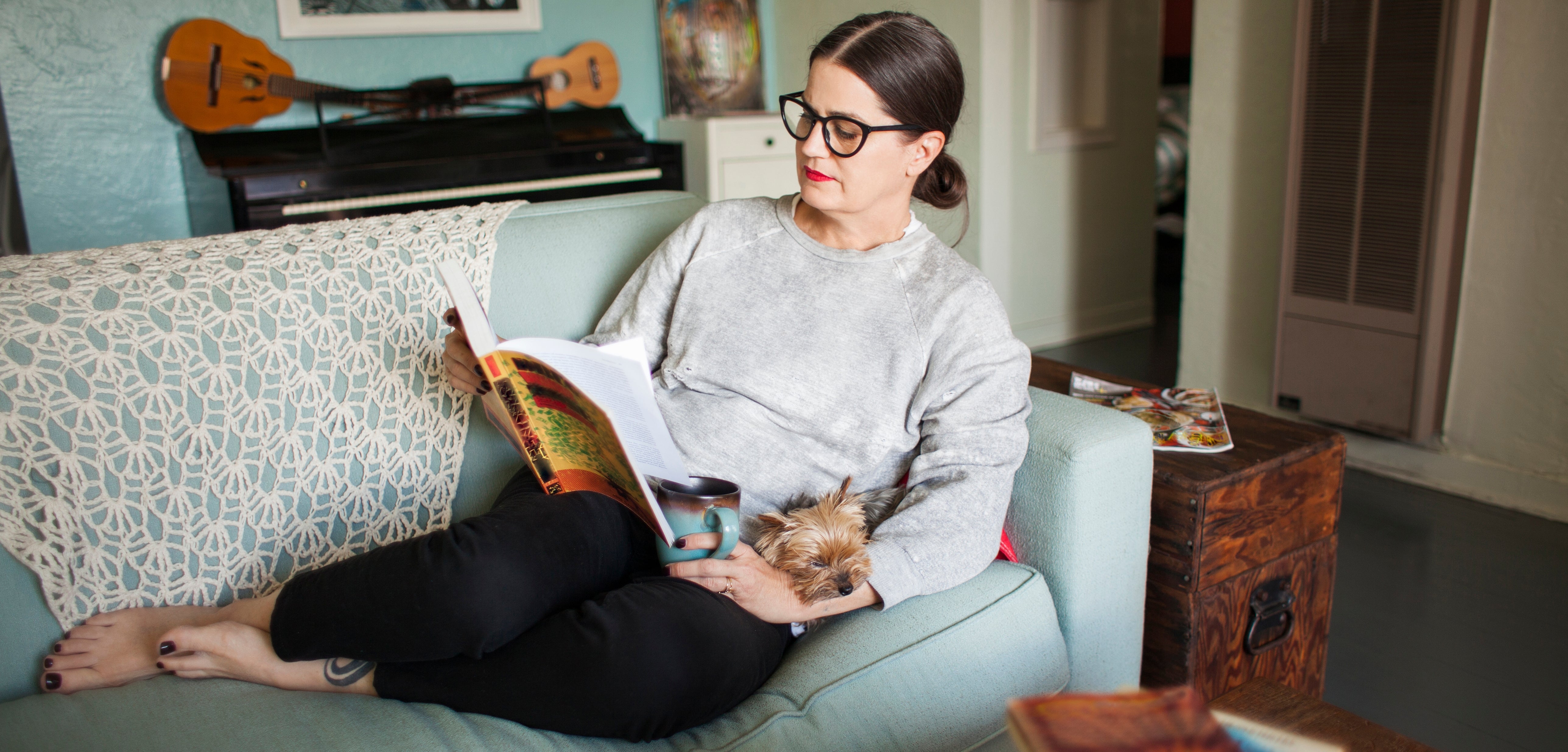 En kvinne sitter på en sofa som leser et magasin, med en liten hund som hviler på fanget. En gitar henger på veggen i bakgrunnen