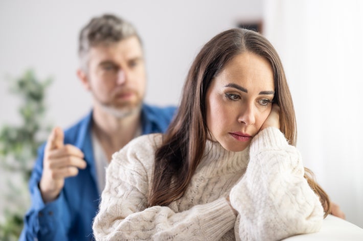 女性が悩んでいる様子で、後ろに男性が指を指して話しているシーン。緊張感が漂う室内の状況。