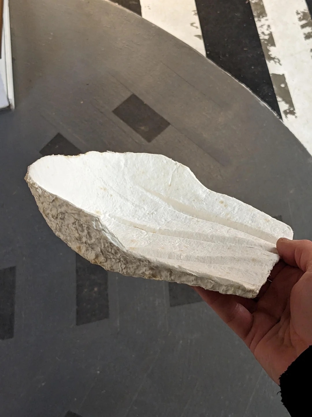 Hand holds a textured, abstract stone-like sculpture with carved grooves, set against a patterned floor background