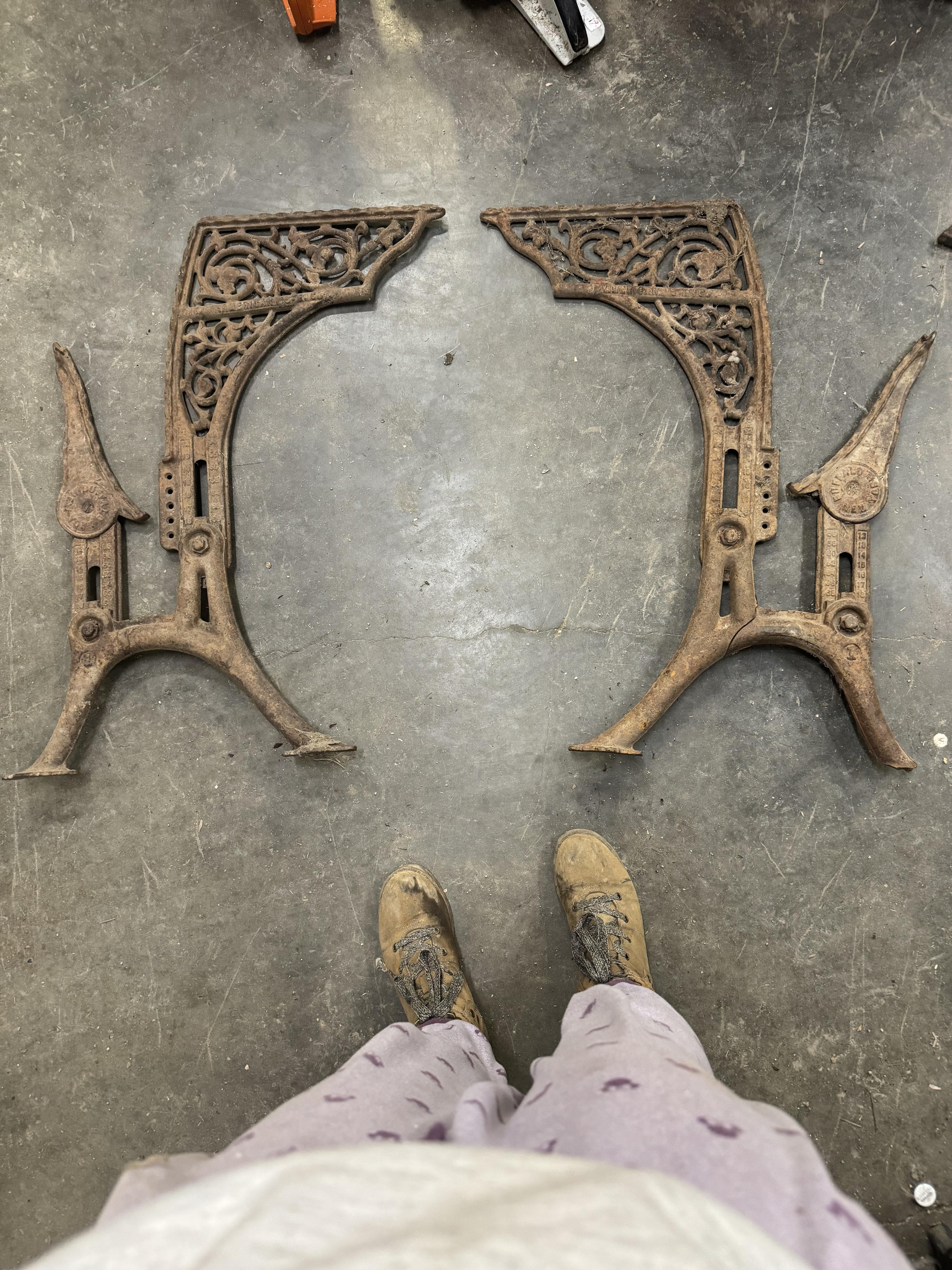 Two ornate, antique, cast iron legs are on a concrete floor next to a person wearing worn boots and patterned pants