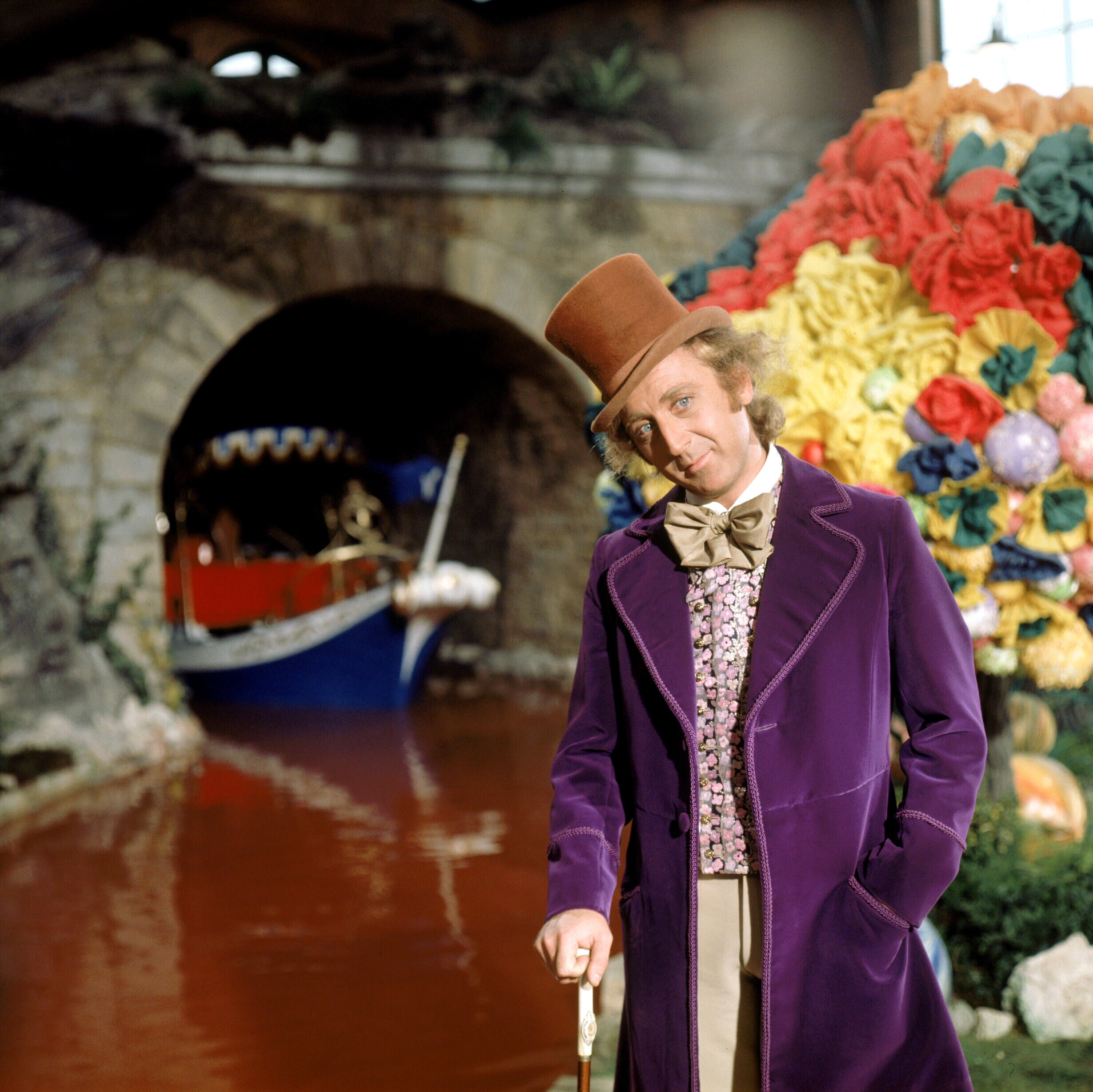 Actor in a whimsical factory setting wearing a velvet coat and top hat, standing by a chocolate river with a boat and colorful candy display