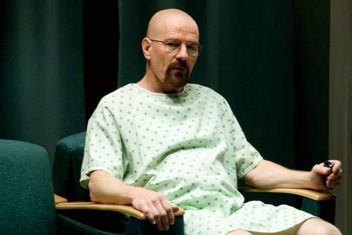 A bald man in a hospital gown sits in a chair holding a remote, appearing deep in thought