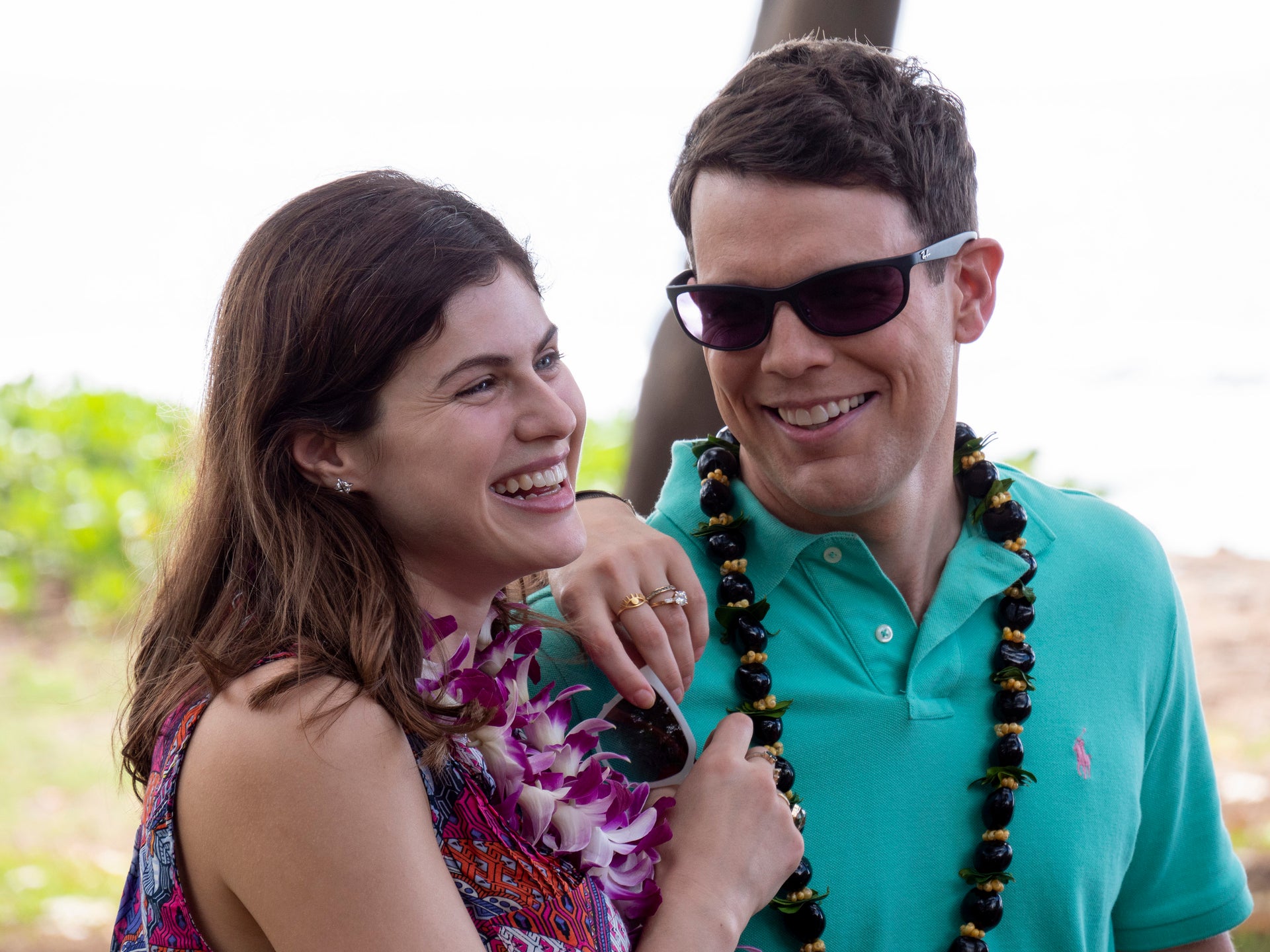Alexandra Daddario and Jake Lacy smiling and wearing leis, outdoors. One wears a floral dress, the other a teal polo and sunglasses