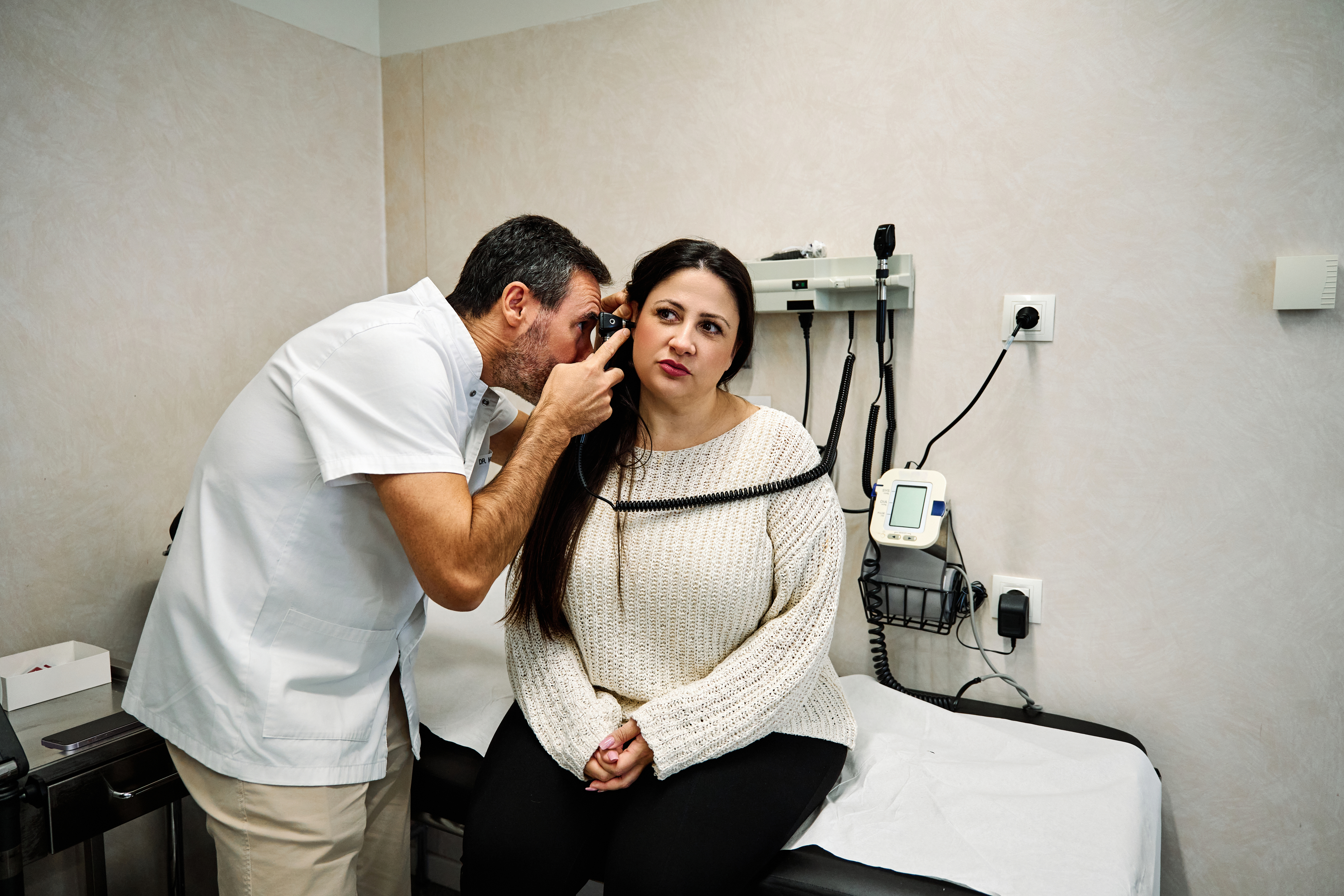 Doctor examining a woman's ear with an otoscope in a medical office. She is sitting on an examination table, wearing a knit sweater