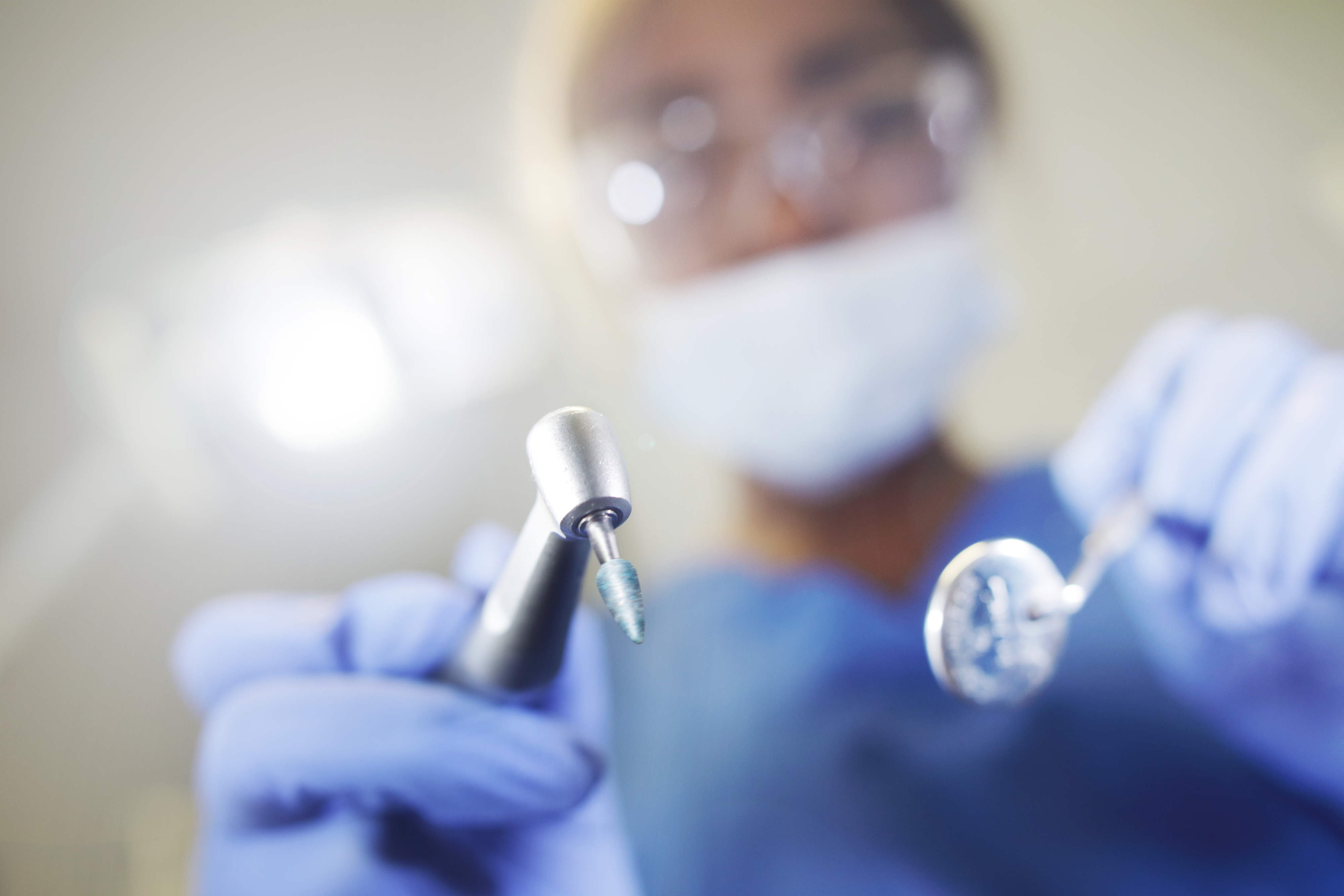 Dentist in mask and gloves holding dental drill and mirror, preparing for a procedure, blurred face and bright light in background
