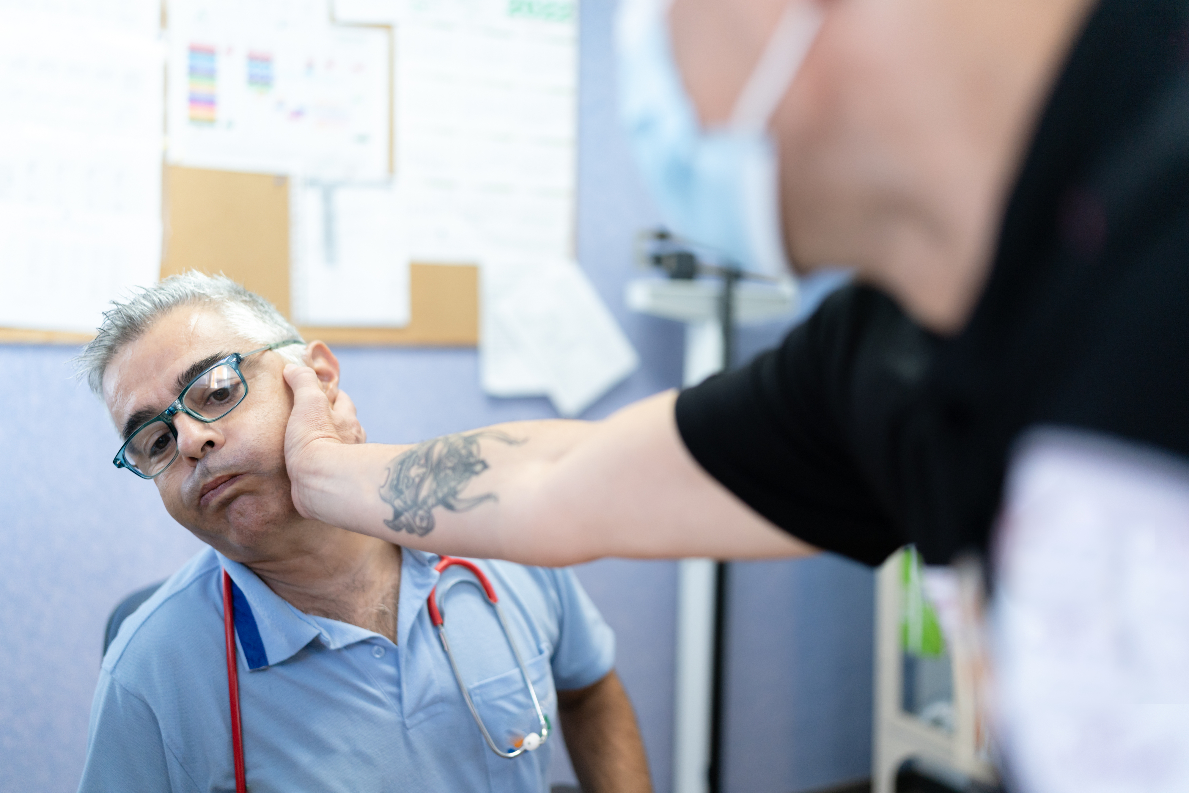A man in scrubs and glasses, with a stethoscope, is being playfully tapped on the cheek by another person. A tattooed arm is visible