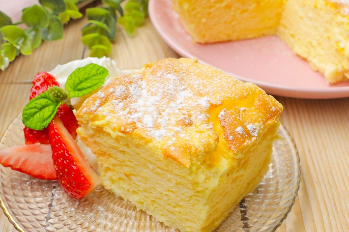 クリームとイチゴが添えられた、ふわふわのケーキの断面が見える。背景には緑の葉がある。