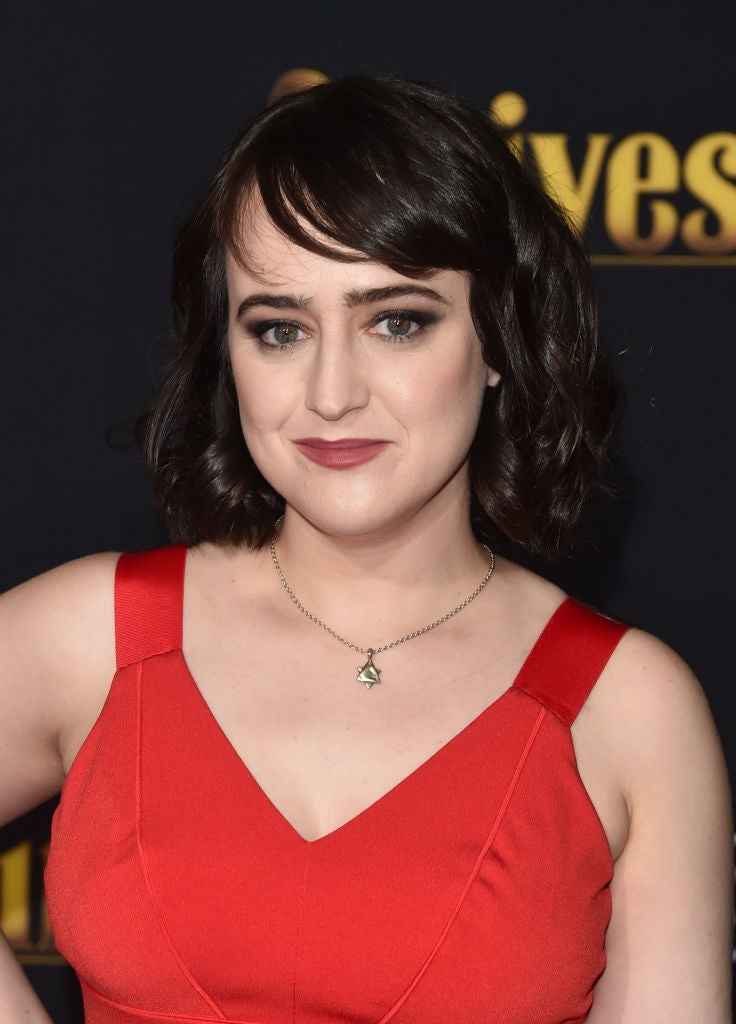 adult mara in a sleeveless red dress with dark hair poses confidently in front of a backdrop with partially visible gold lettering