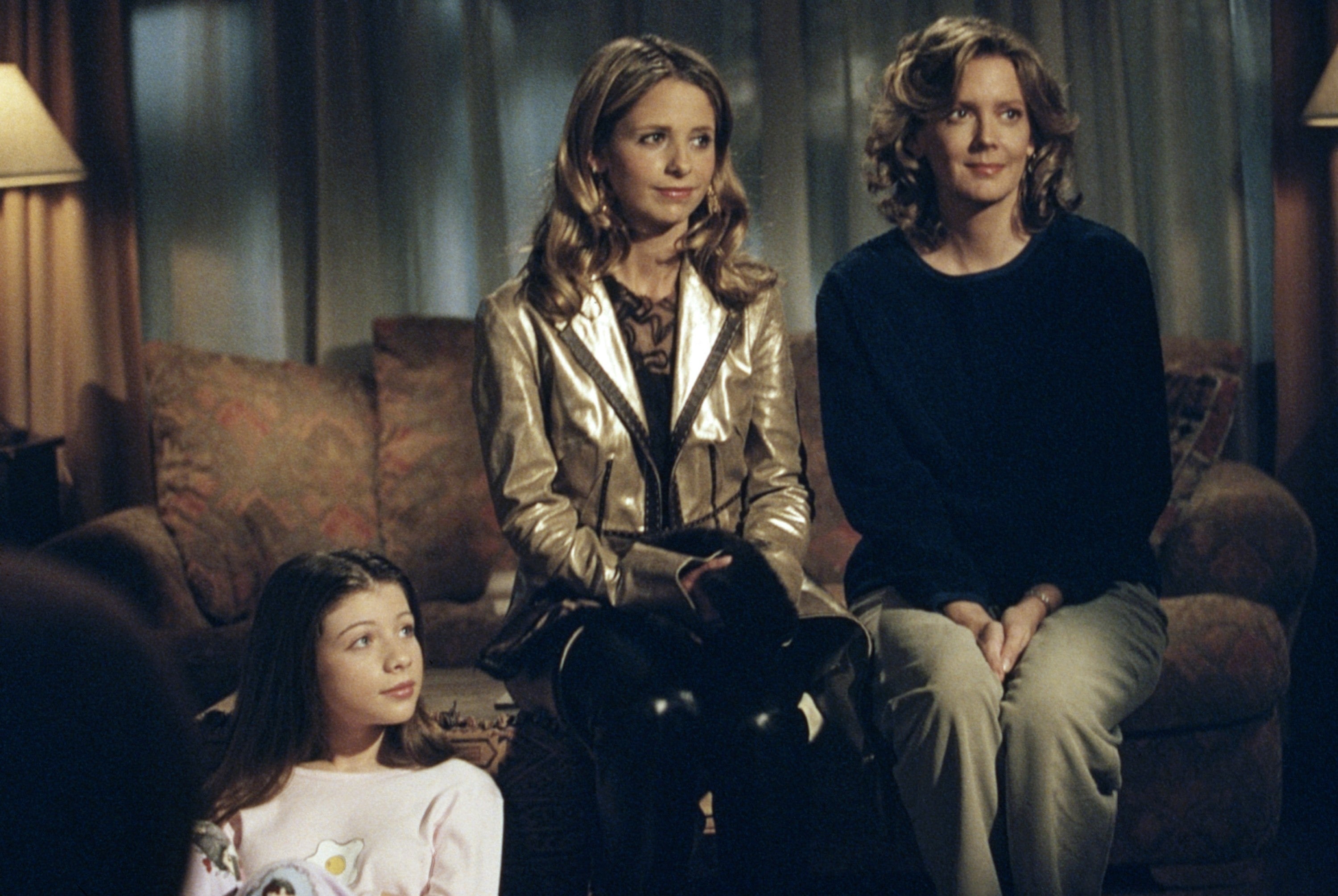 Three people are sitting indoors on a sofa. The center person wears a shiny jacket, while the others wear casual tops. They appear relaxed