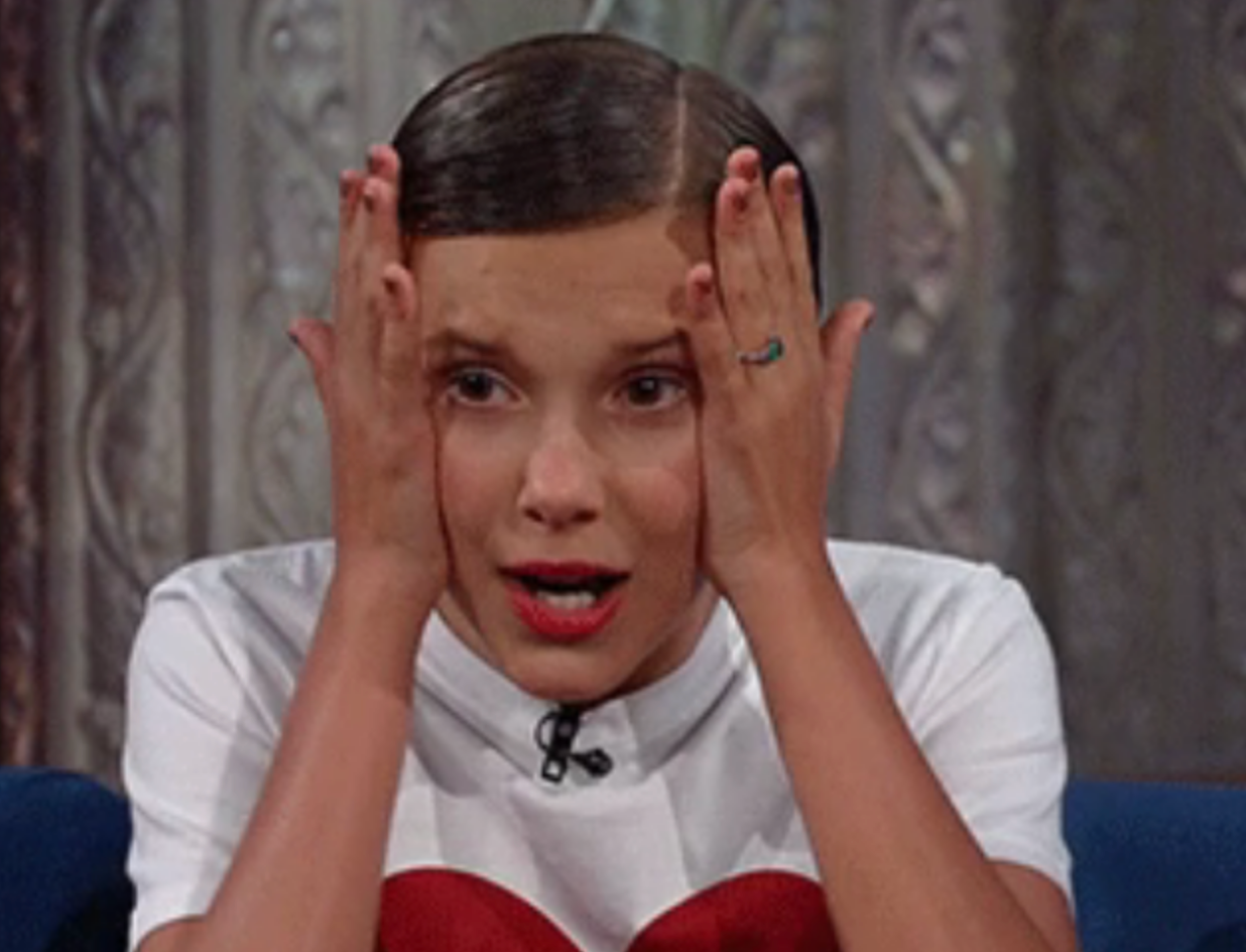 Person holding face with both hands, wearing a white shirt, looking surprised or amused during an interview on a TV show