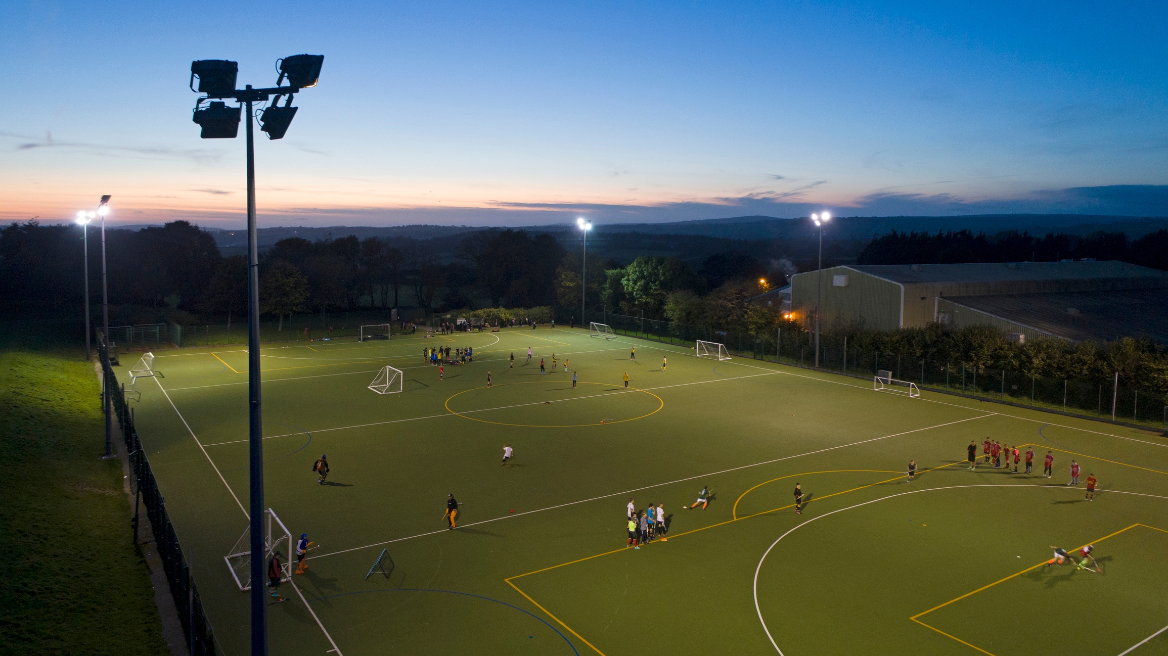 A sports field at dusk with several groups playing soccer under bright floodlights. Hills and buildings are visible in the background