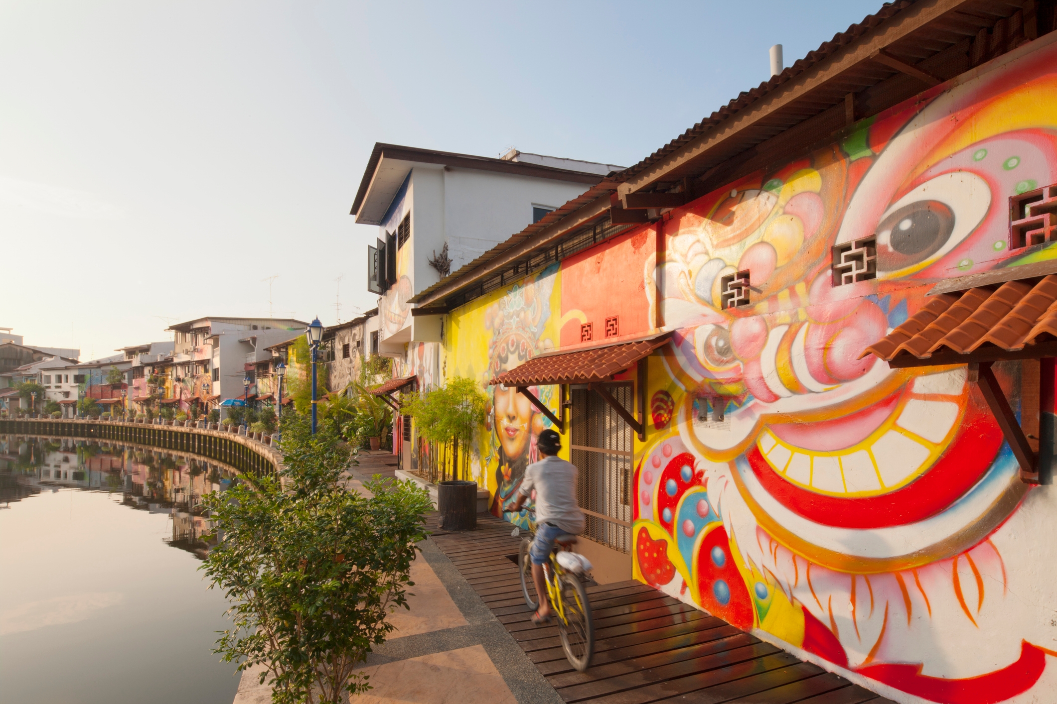 Person on bicycle rides past vibrant dragon mural along river in urban setting, with colorful buildings and clear sky reflecting on water