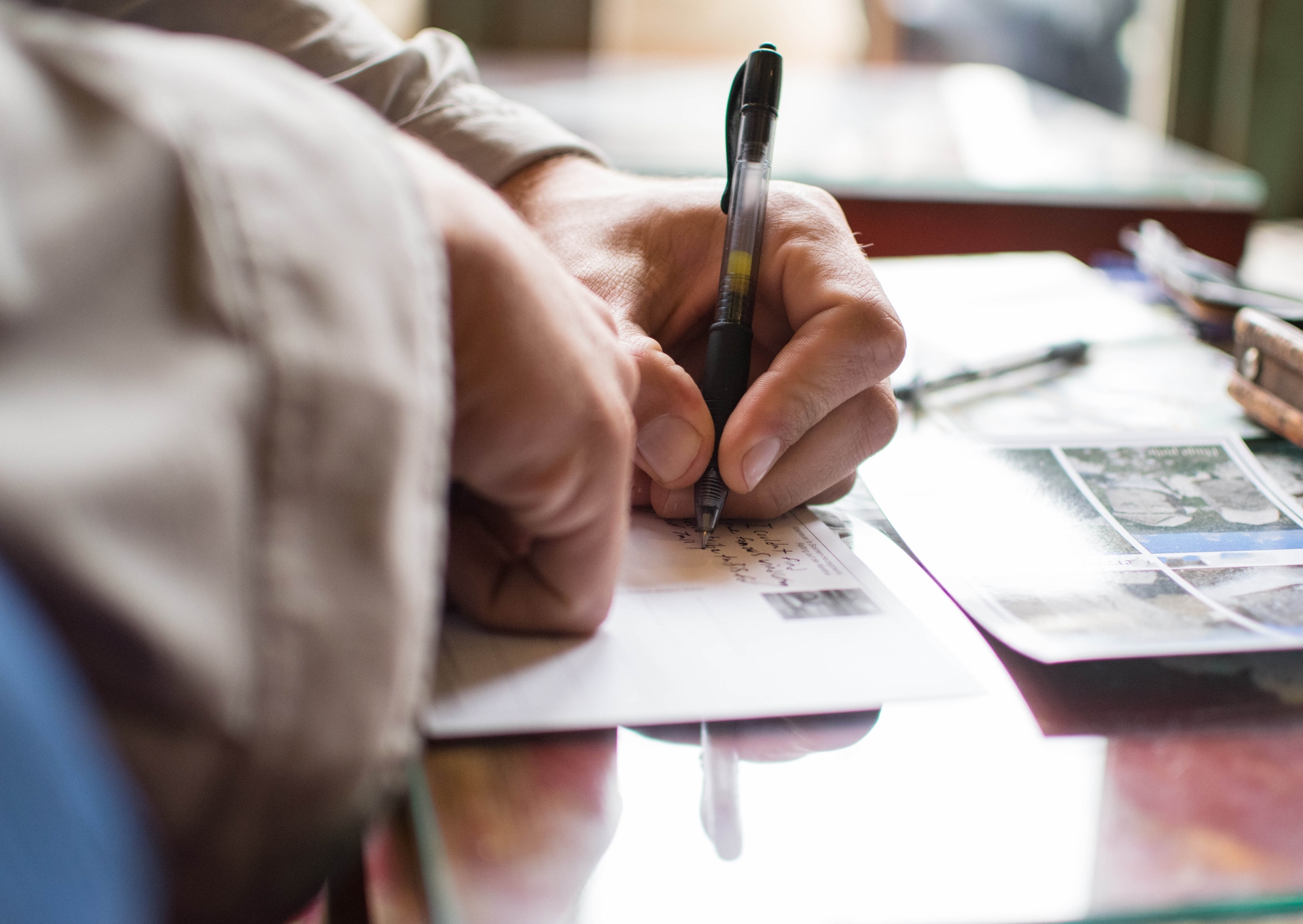 Person writing on a postcard at a table with pens and papers nearby