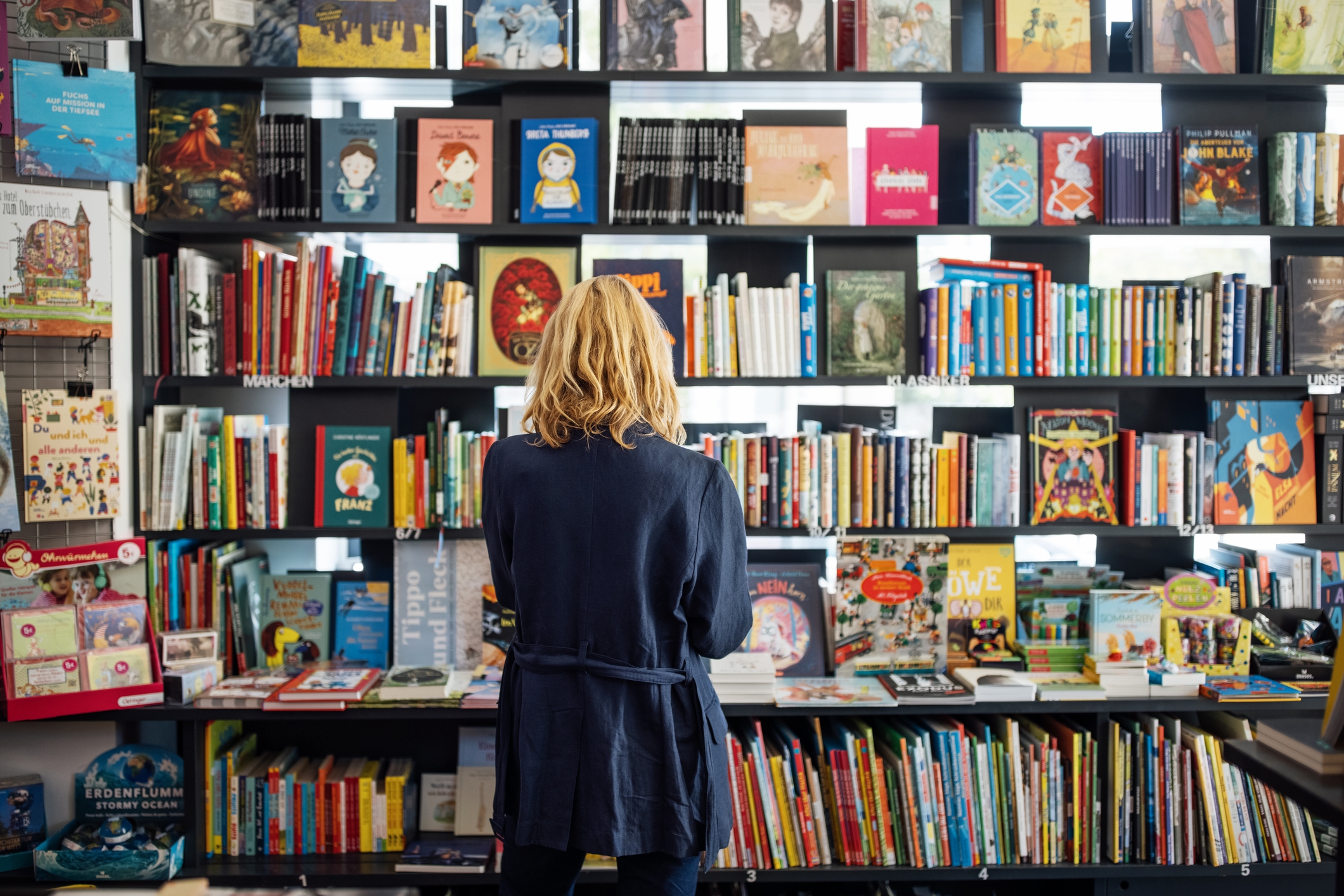 Person with shoulder-length hair browsing a colorful and diverse collection of books in a bookstore, standing with their back to the camera