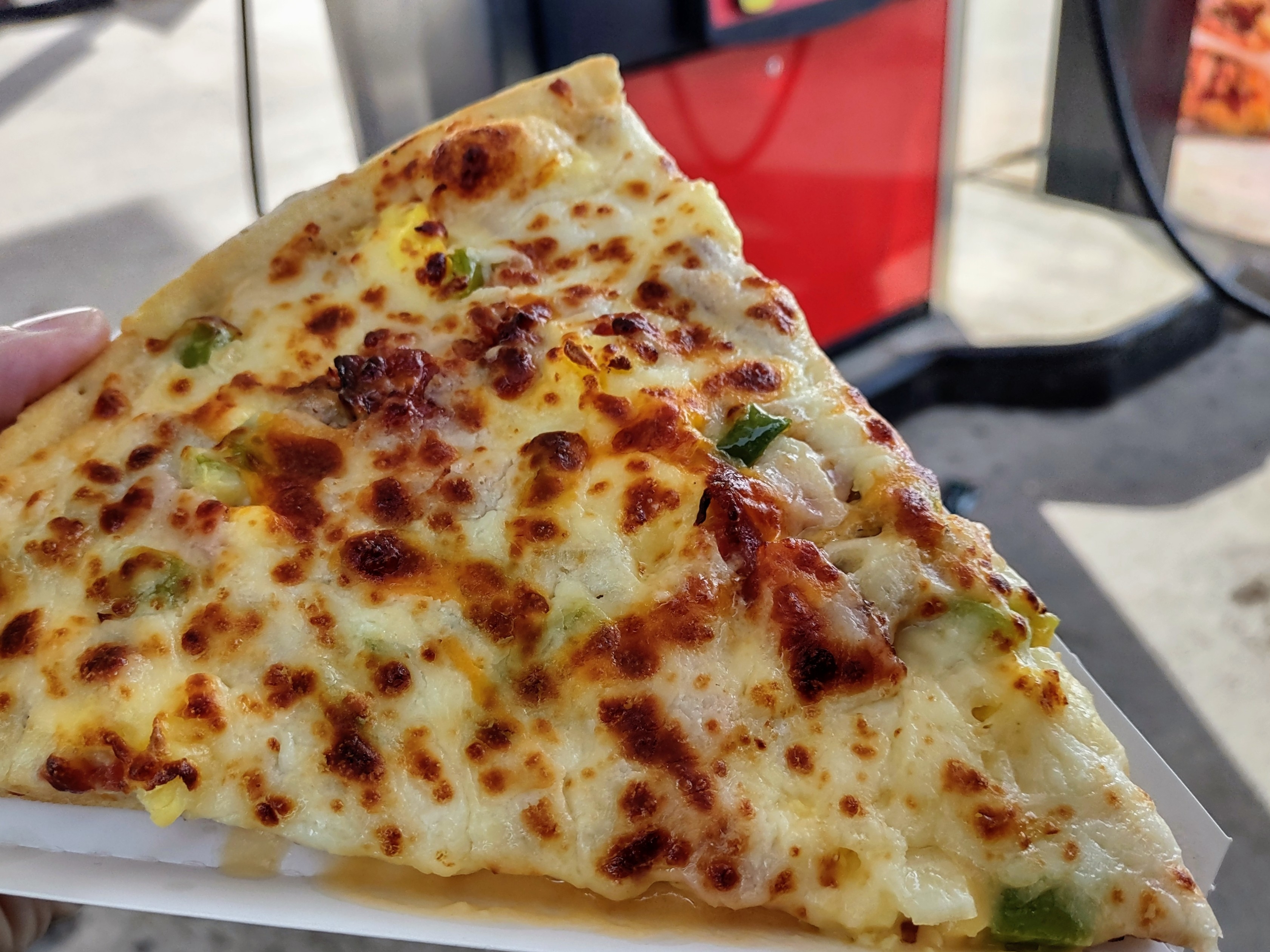 A slice of pizza with melted cheese and vegetable toppings, held outdoors near a gas station