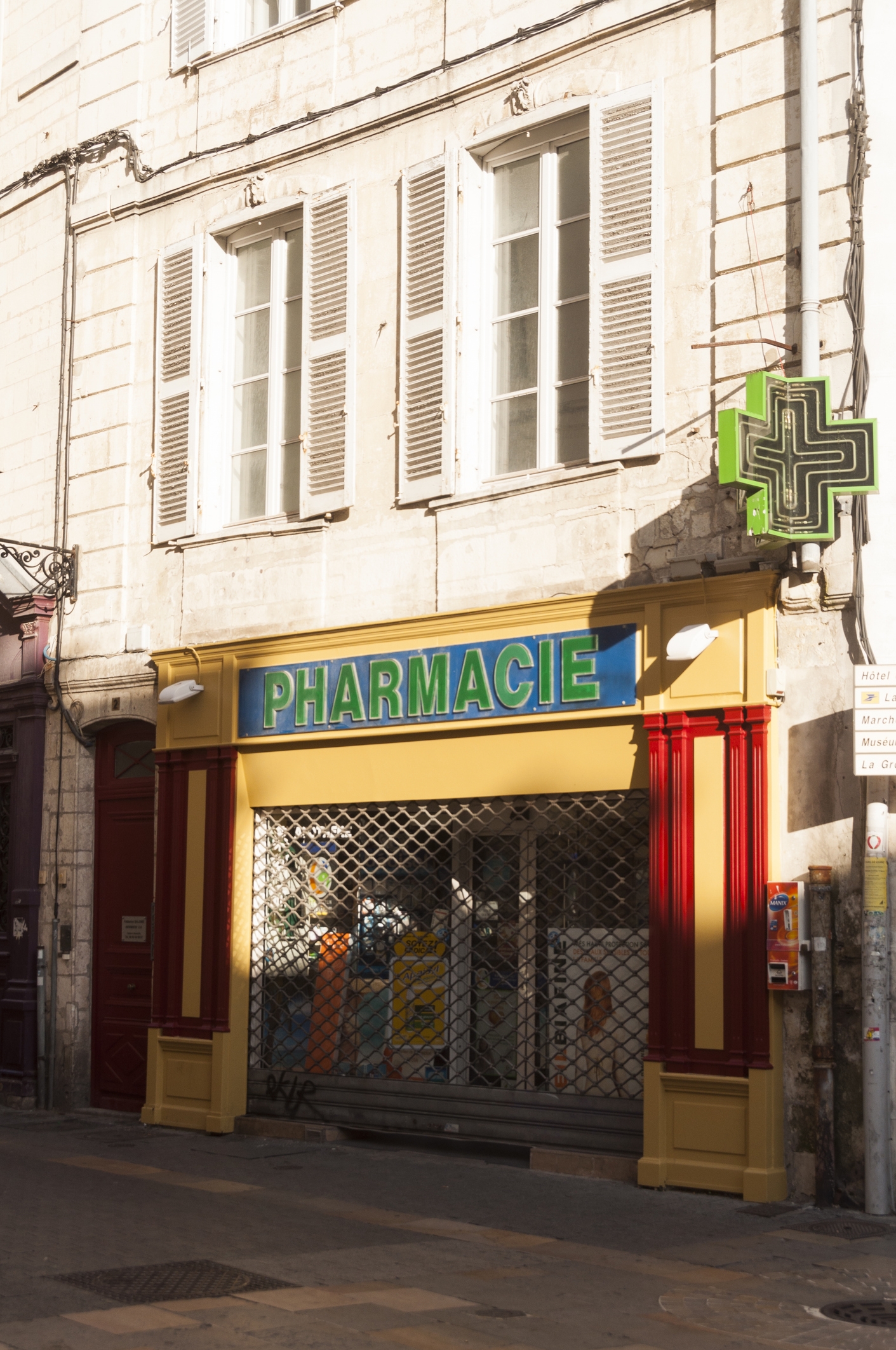 Storefront of a closed pharmacy with a metal gate down; a green cross symbol for pharmacies in France is visible above