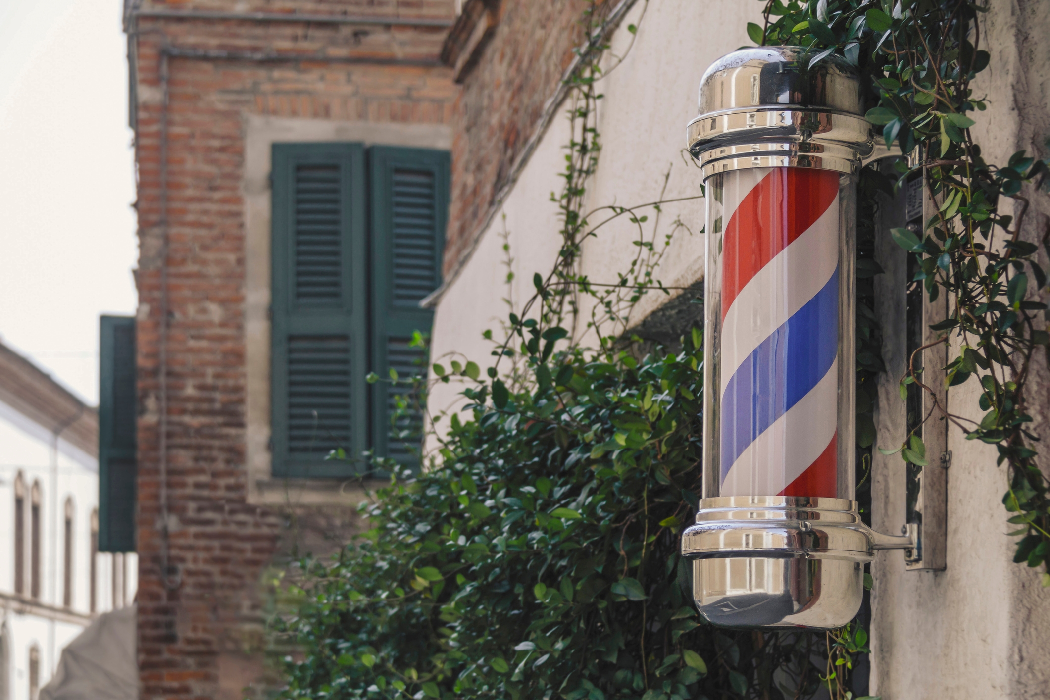 Barber pole with rotating red, white, and blue stripes mounted on an ivy-covered wall in a historic-looking neighborhood