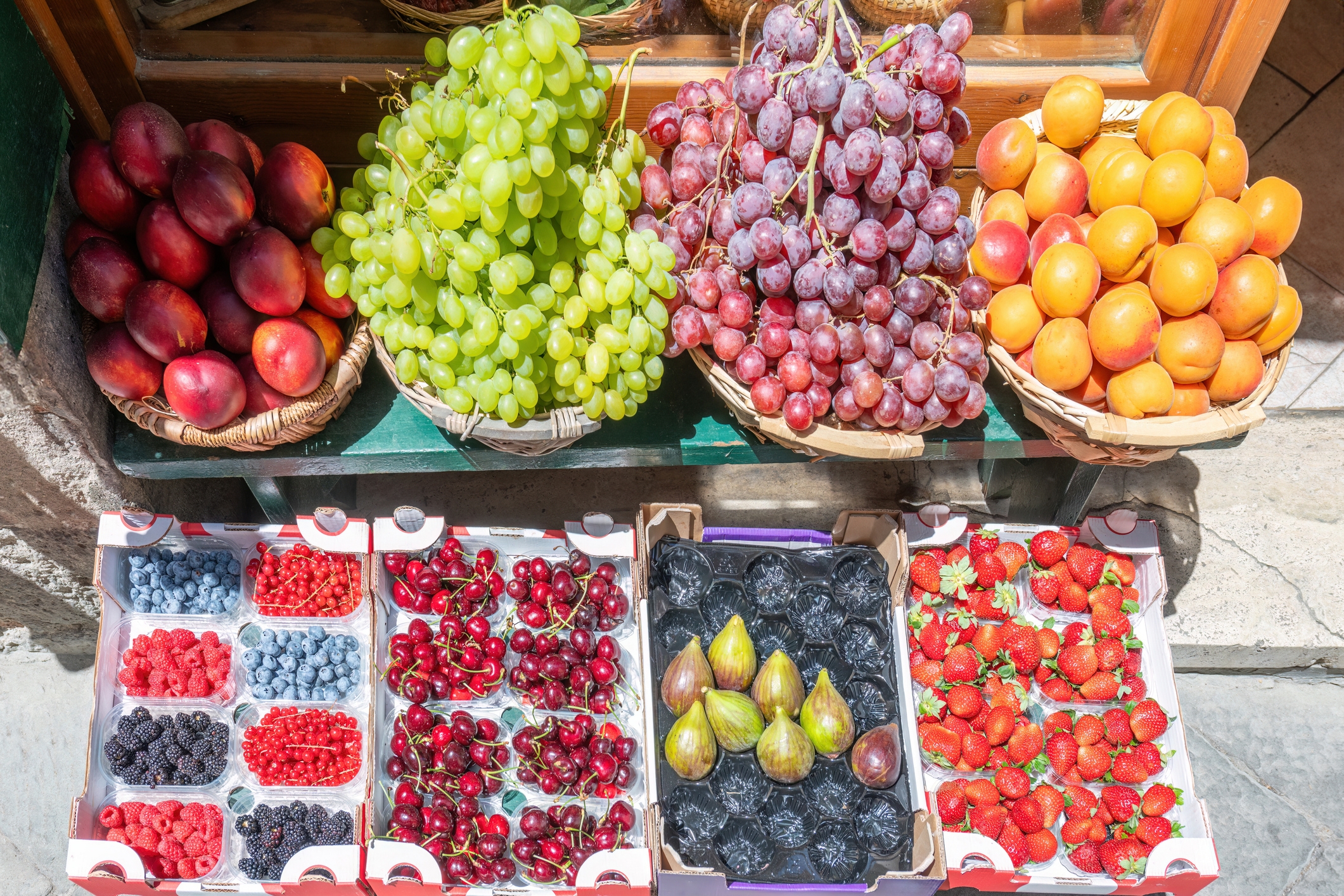 Assorted fruits displayed in baskets and boxes, including grapes, peaches, figs, strawberries, blueberries, and cherries at a market