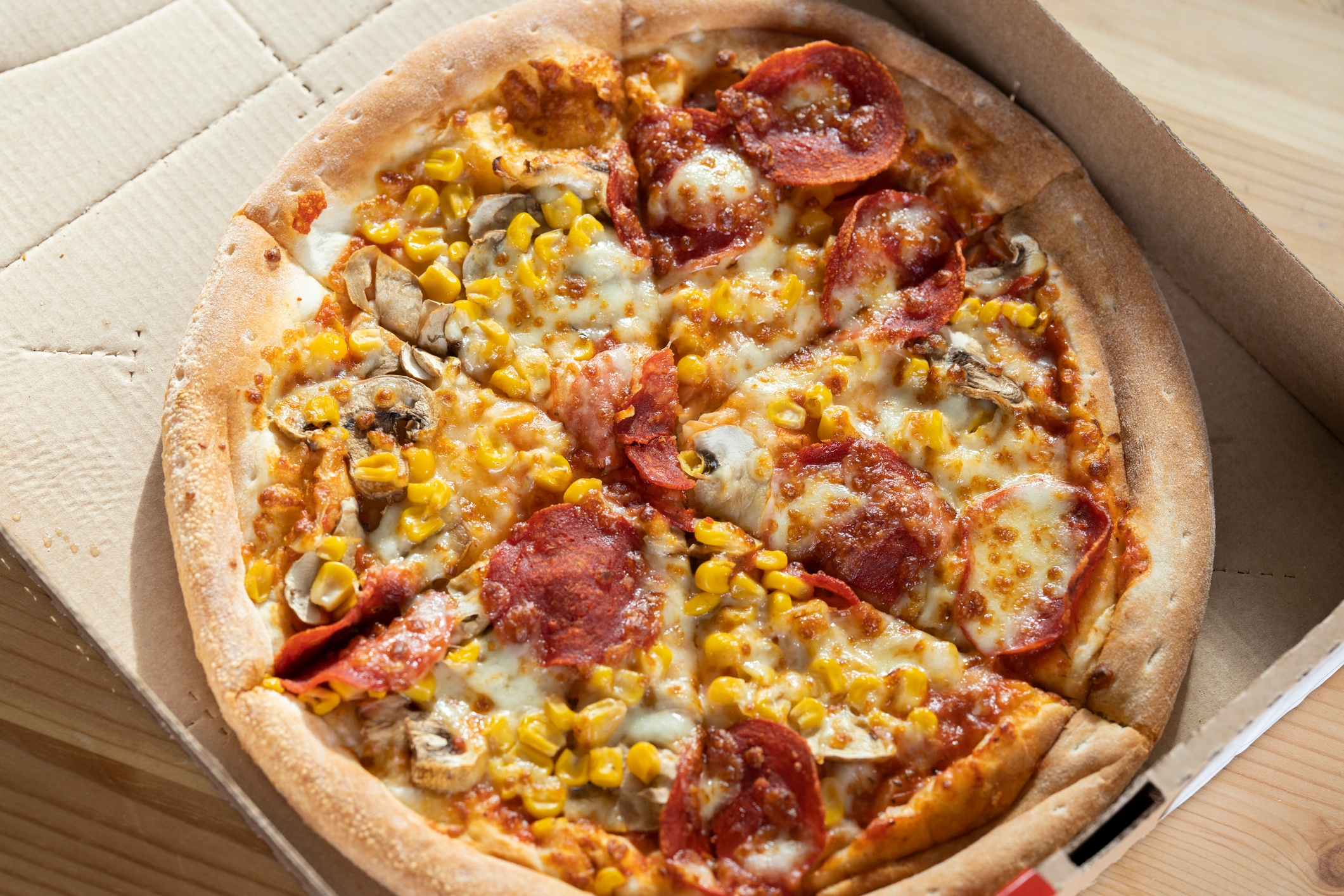 Pizza with pepperoni, corn, mushrooms, and melted cheese in an open box on a wooden table