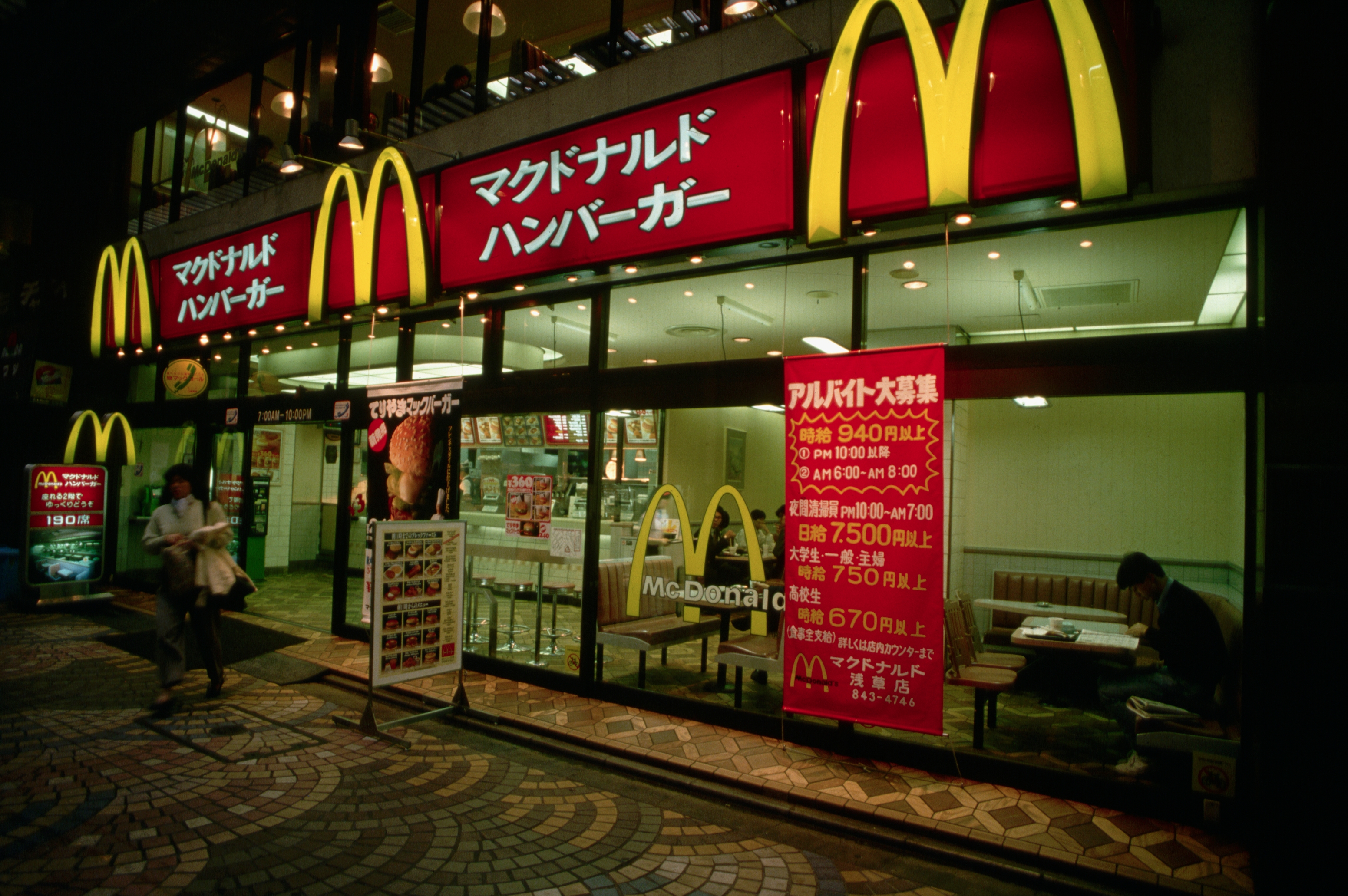 Street view of a McDonald's in Japan with people walking by and text in Japanese on a large sign outside