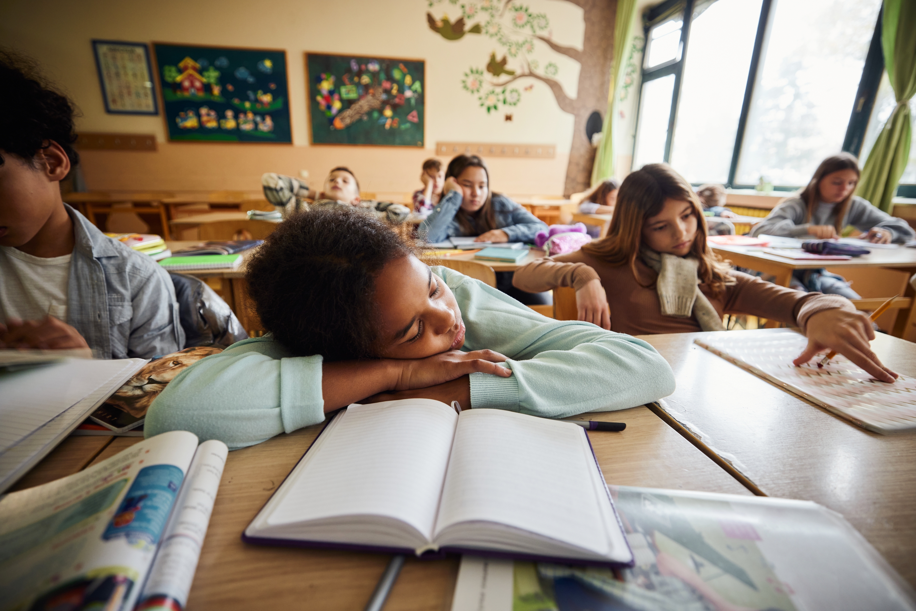 Et klasserom med elever som sitter på pulter. En student i forgrunnen har sovnet med en bok åpen foran dem
