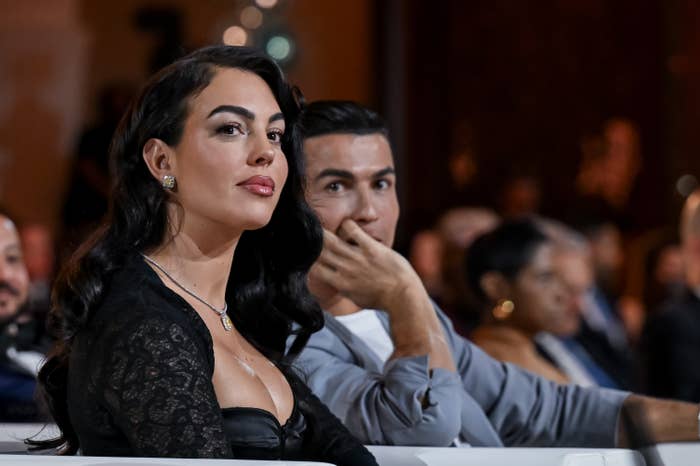 A woman in a lace dress and a man in a casual shirt sit attentively at a formal event, surrounded by other attendees