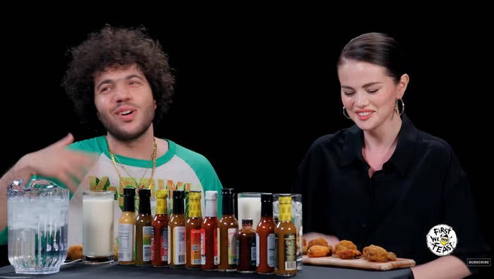 Two individuals are seated at a table with various hot sauces and chicken wings in front of them during a televised interview or challenge show