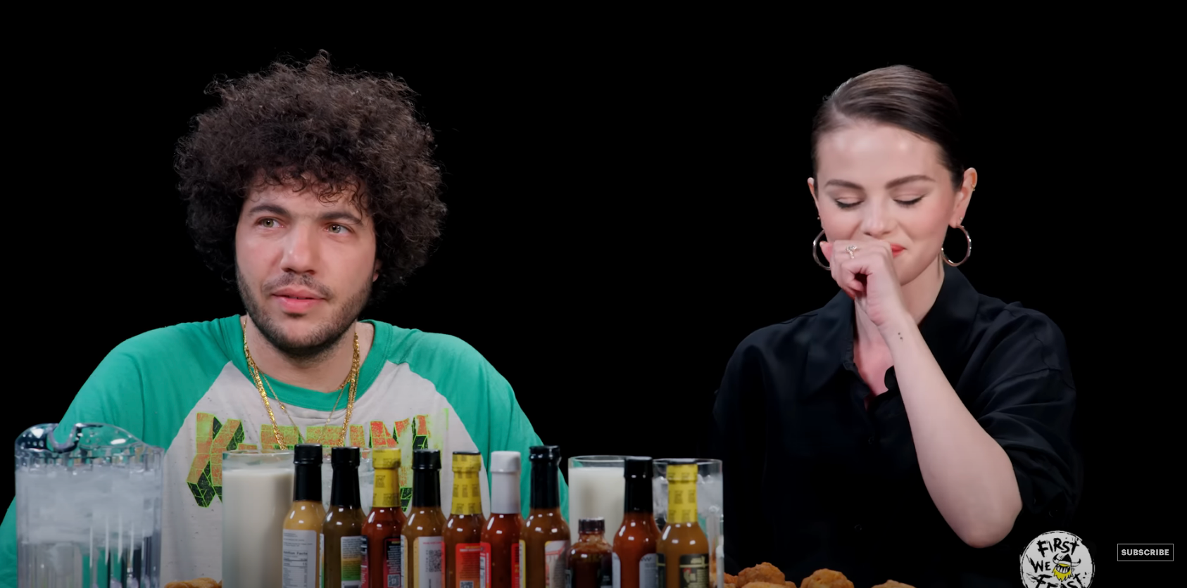 Two individuals participate in an interview show with hot sauces on a table; one looks calm, while the other appears to be reacting to spice