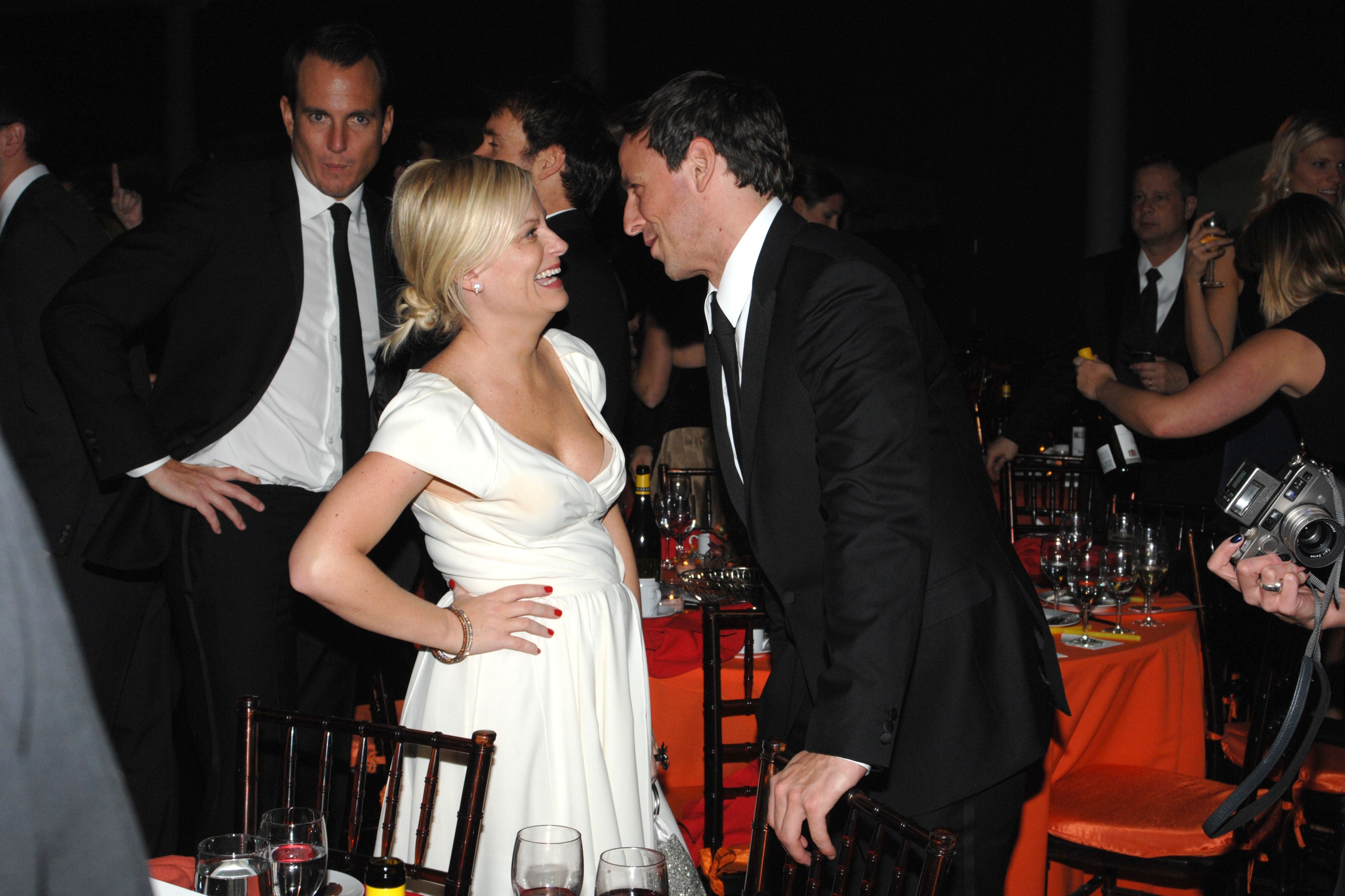 Seth Meyers leaning in to say something to Amy Poehler, who laughs. Will Arnett stands menacingly in the background, hand on hip