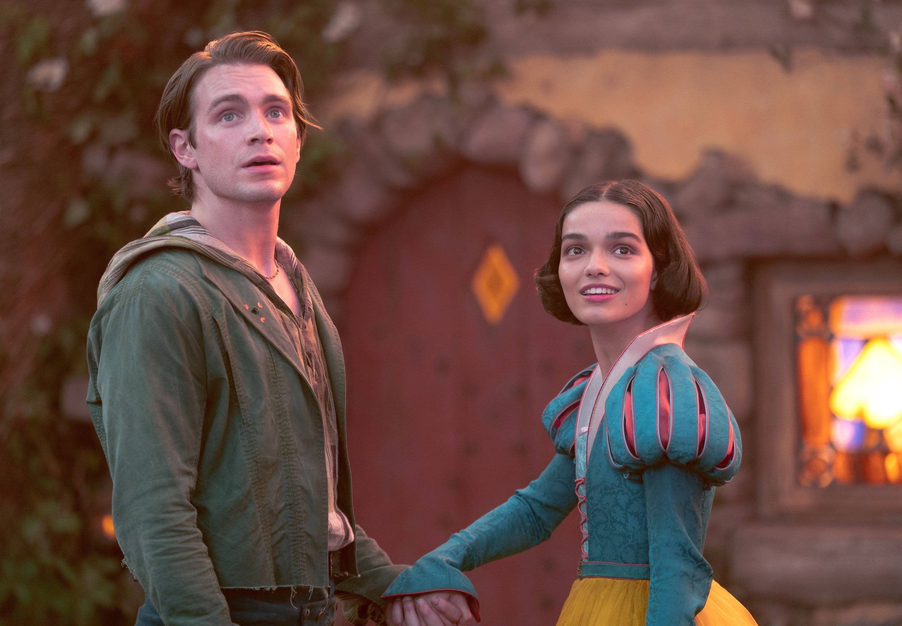 Two actors in fantasy attire hold hands, with one in a hooded outfit and the other in a costume resembling Snow White. Background features a cottage