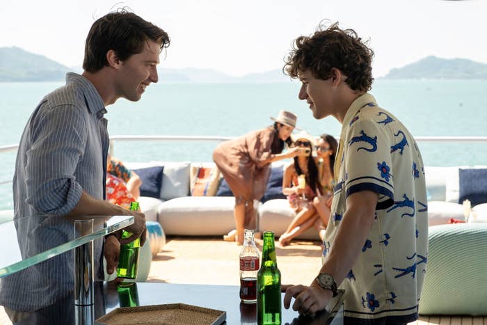 Patrick Schwarzenegger and Sam Nivola talking on a yacht with drinks in hand; guests relax in the background, and a woman in a hat takes a photo