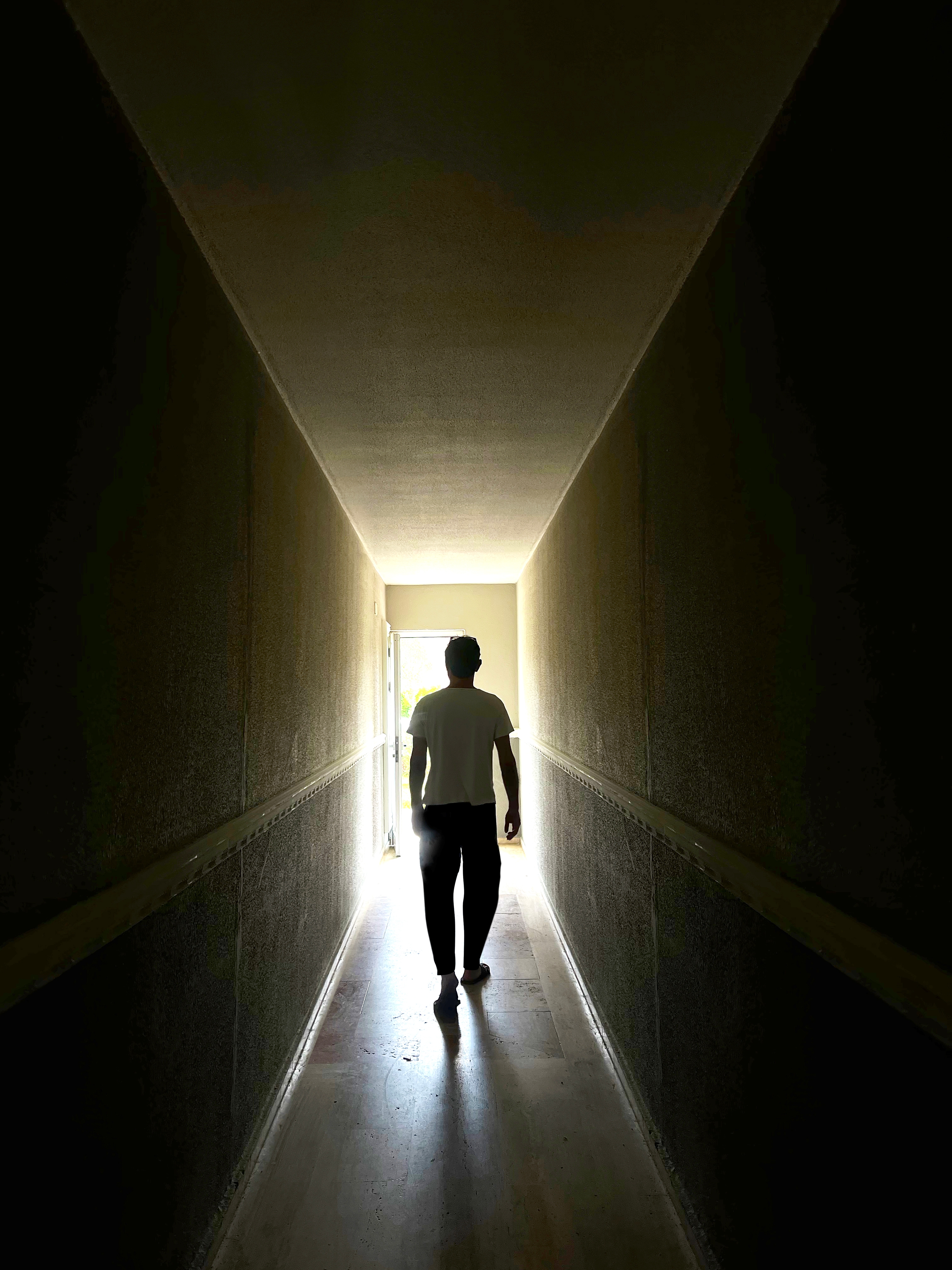 Silhouette of a person walking down a narrow hallway towards a bright exit