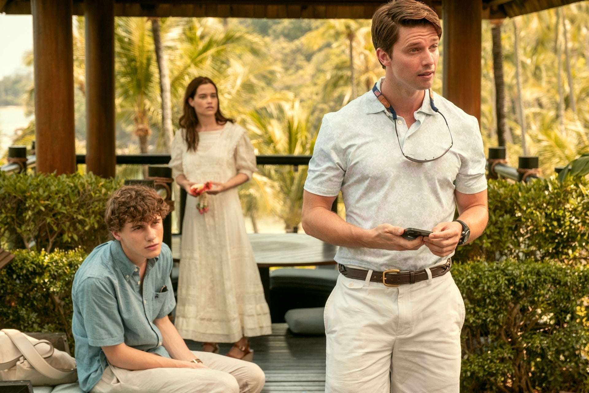 Three people in a tropical setting, one sitting and two standing. The person on the right wears a polo and sunglasses around the neck
