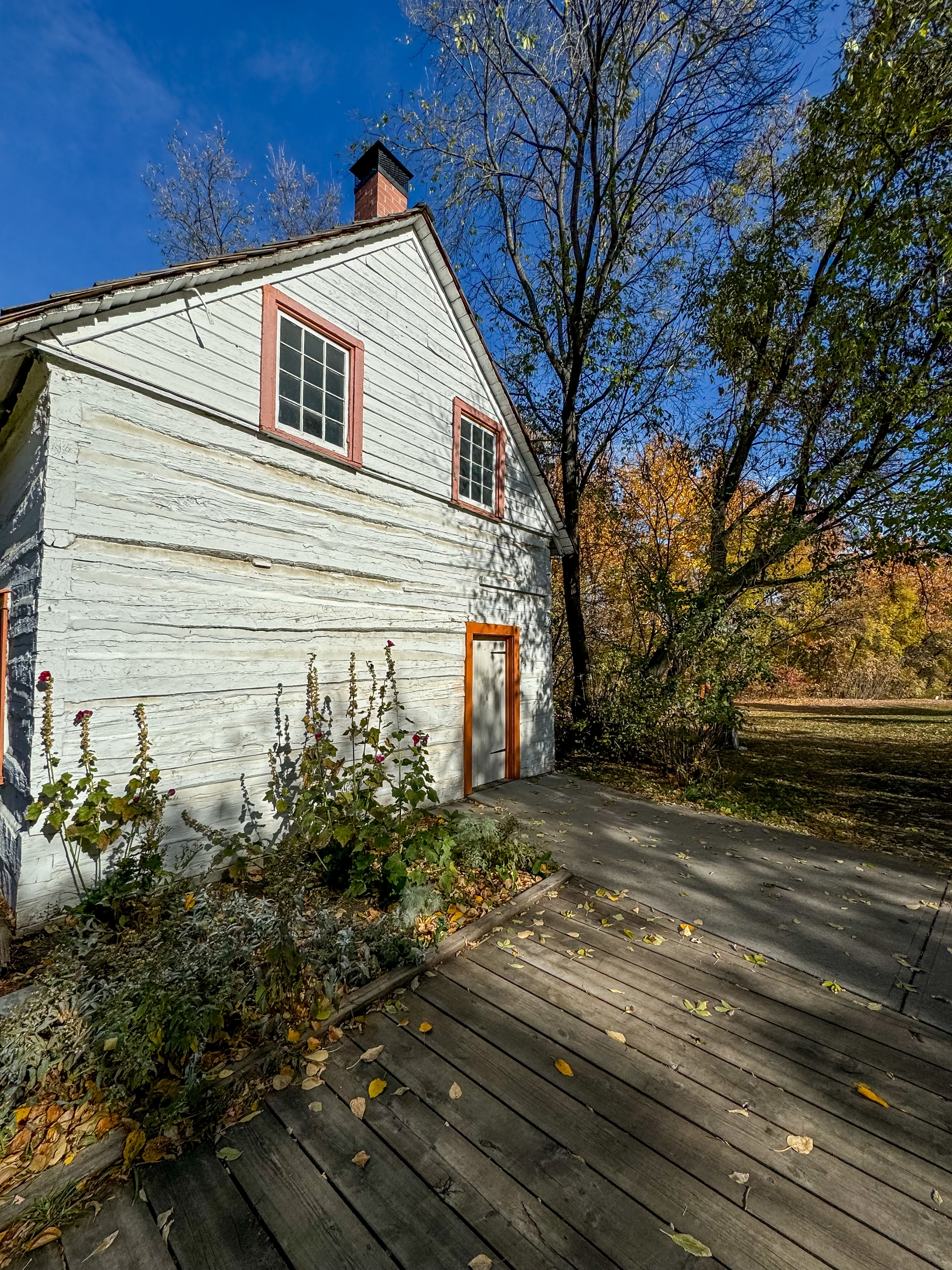 A rustic wooden house with a chimney, surrounded by autumn trees and a garden bed. A wooden deck leads to the door
