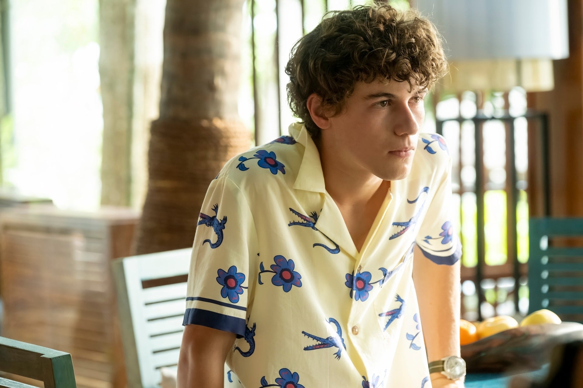 Sam Nivola in floral-patterned shirt leans on a table, looking contemplative, in a room with blurred background elements