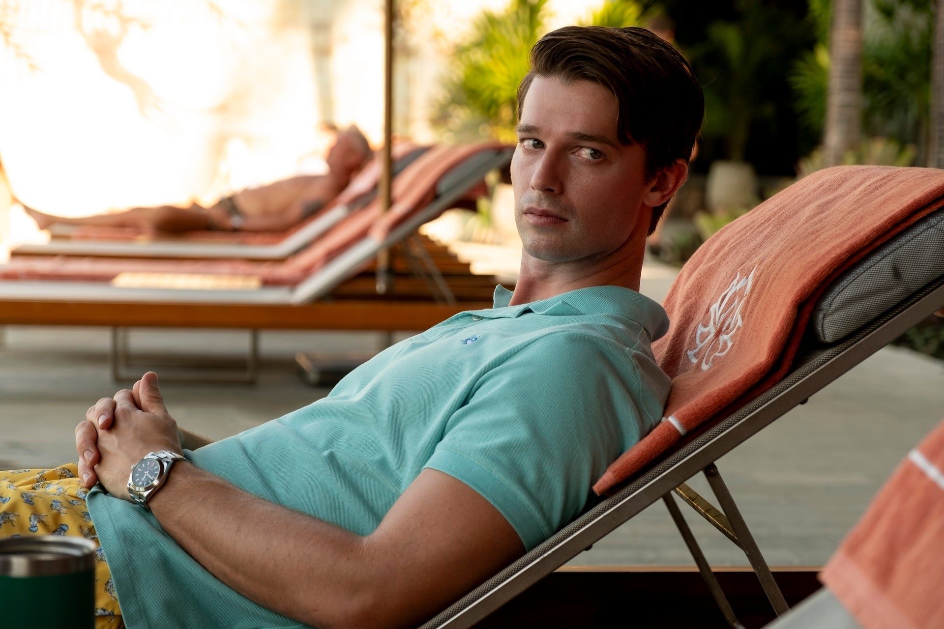 Patrick Schwarzenegger in casual summer attire lounges on a poolside chair, looking towards the camera