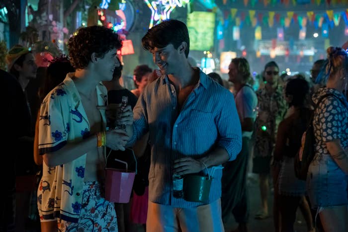 Sam Nivola and Patrick Schwarzenegger at a lively party, one in a patterned shirt, the other in a striped shirt, holding drinks and enjoying a conversation in a festive atmosphere