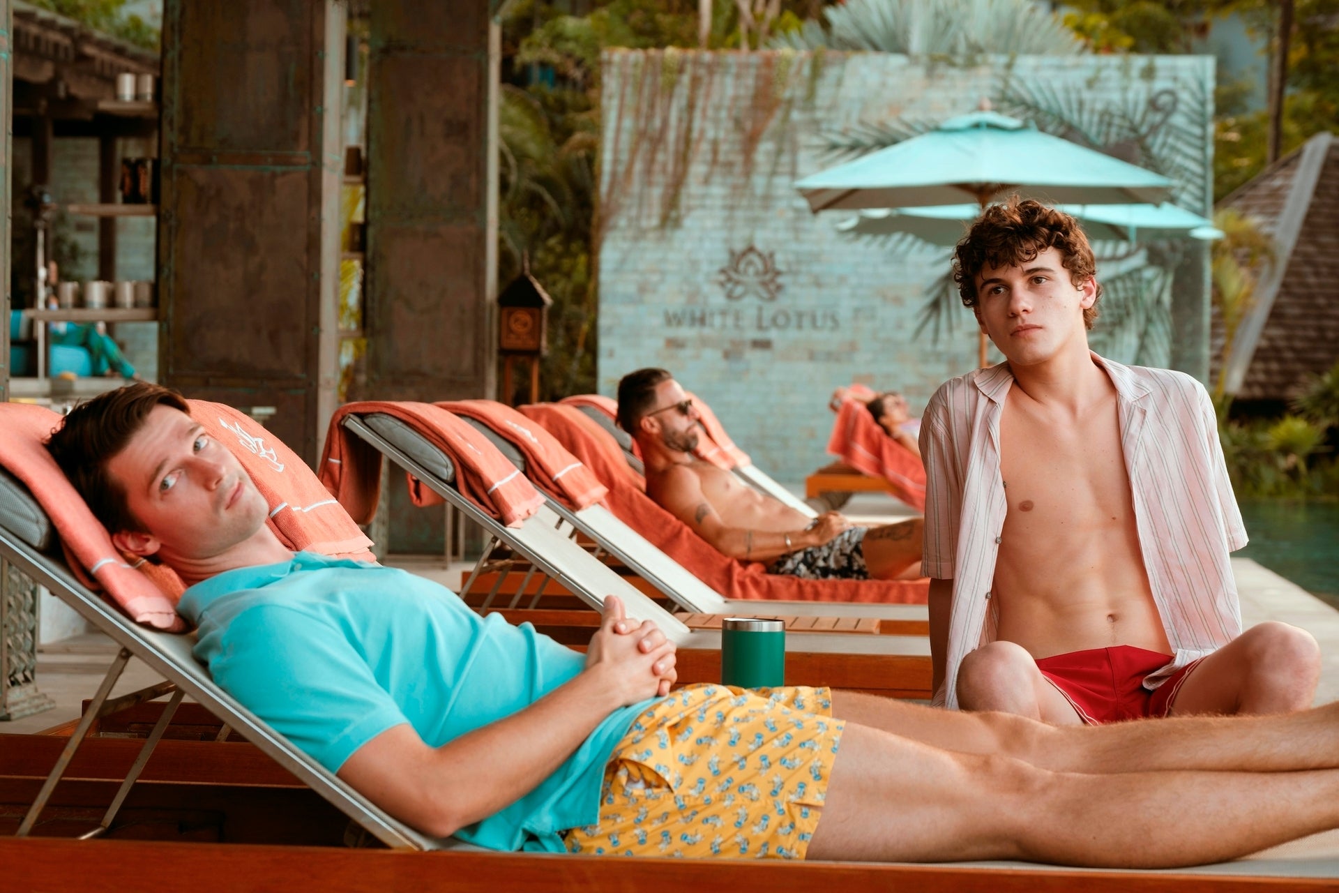 Patrick Schwarzenegger and Sam Nivola relax on lounge chairs by a pool. Two in the foreground have casual, summer attire, with patterned shorts and open shirts
