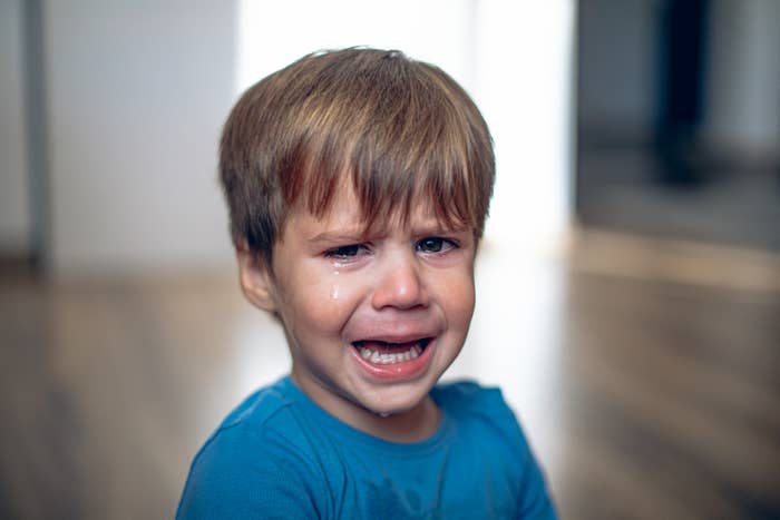 A young child with tears on their face has a distressed expression, wearing a simple shirt, in an indoor setting with a blurred background
