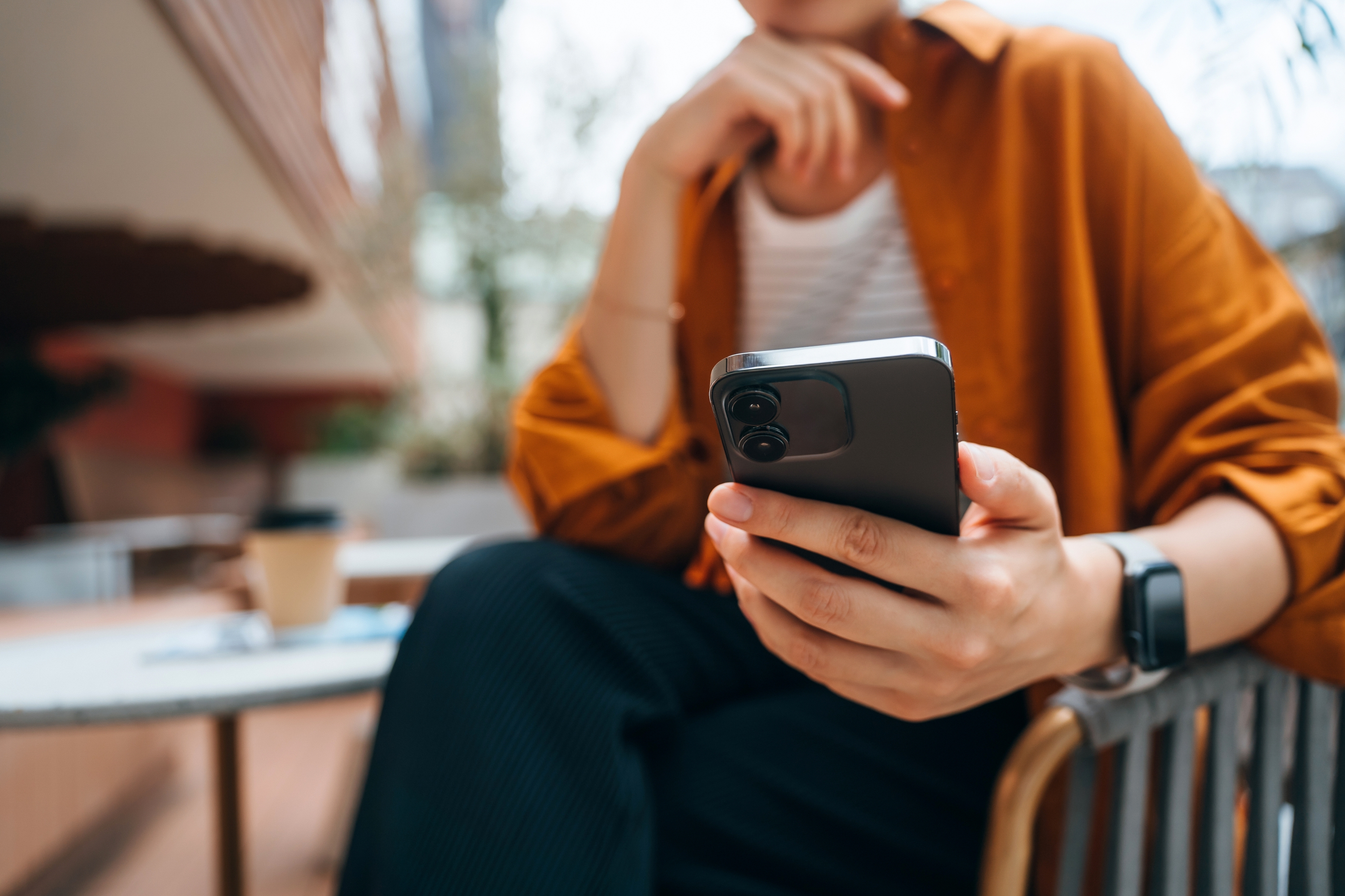 Person som sitter utendørs, ser på telefonen sin, har på seg en uformell skjorte og ser på. En kaffekopp er på bordet ved siden av dem