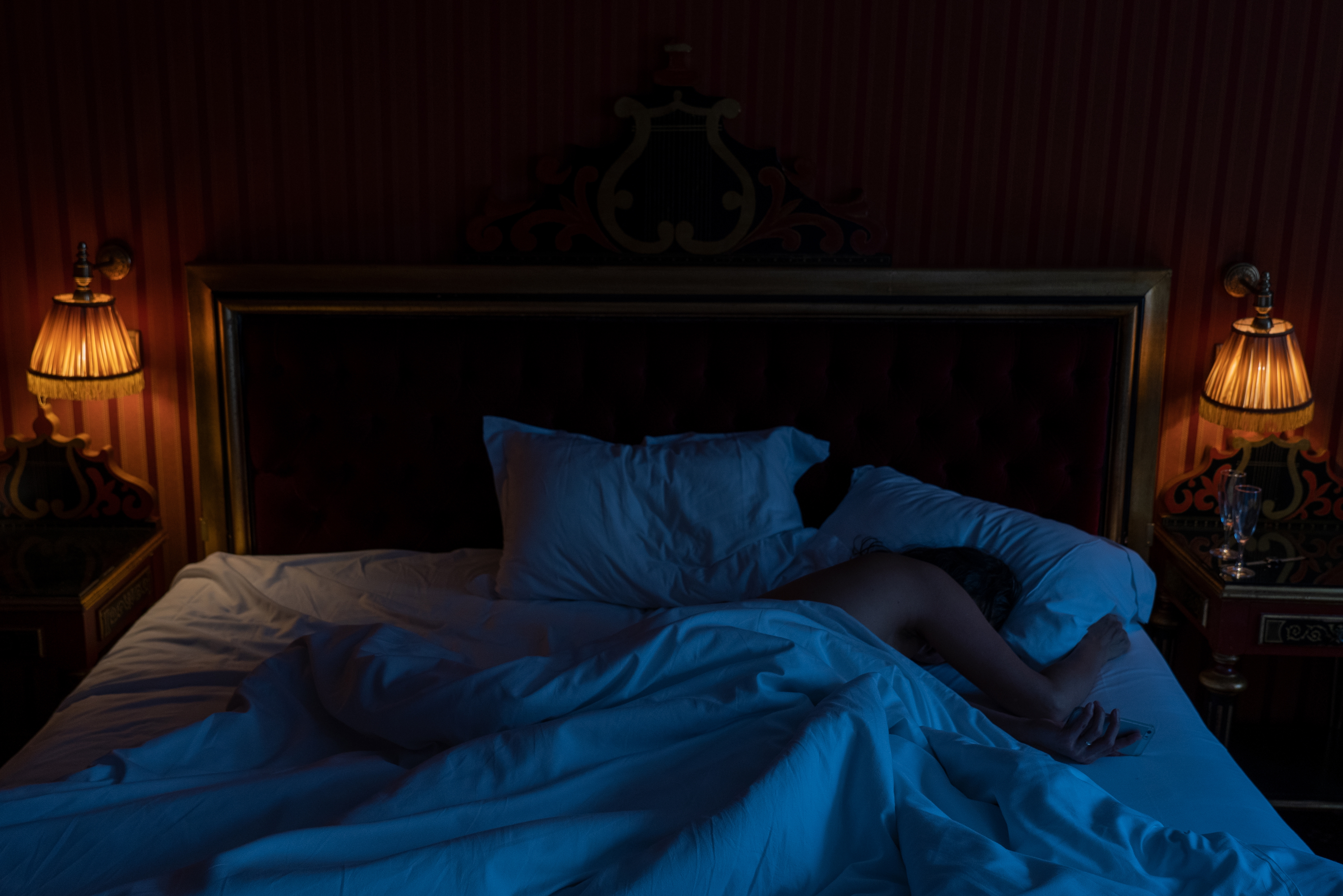 Personen som sover i et svakt opplyst rom, dekket av et teppe på en stor seng med utsmykkede lamper på hver side