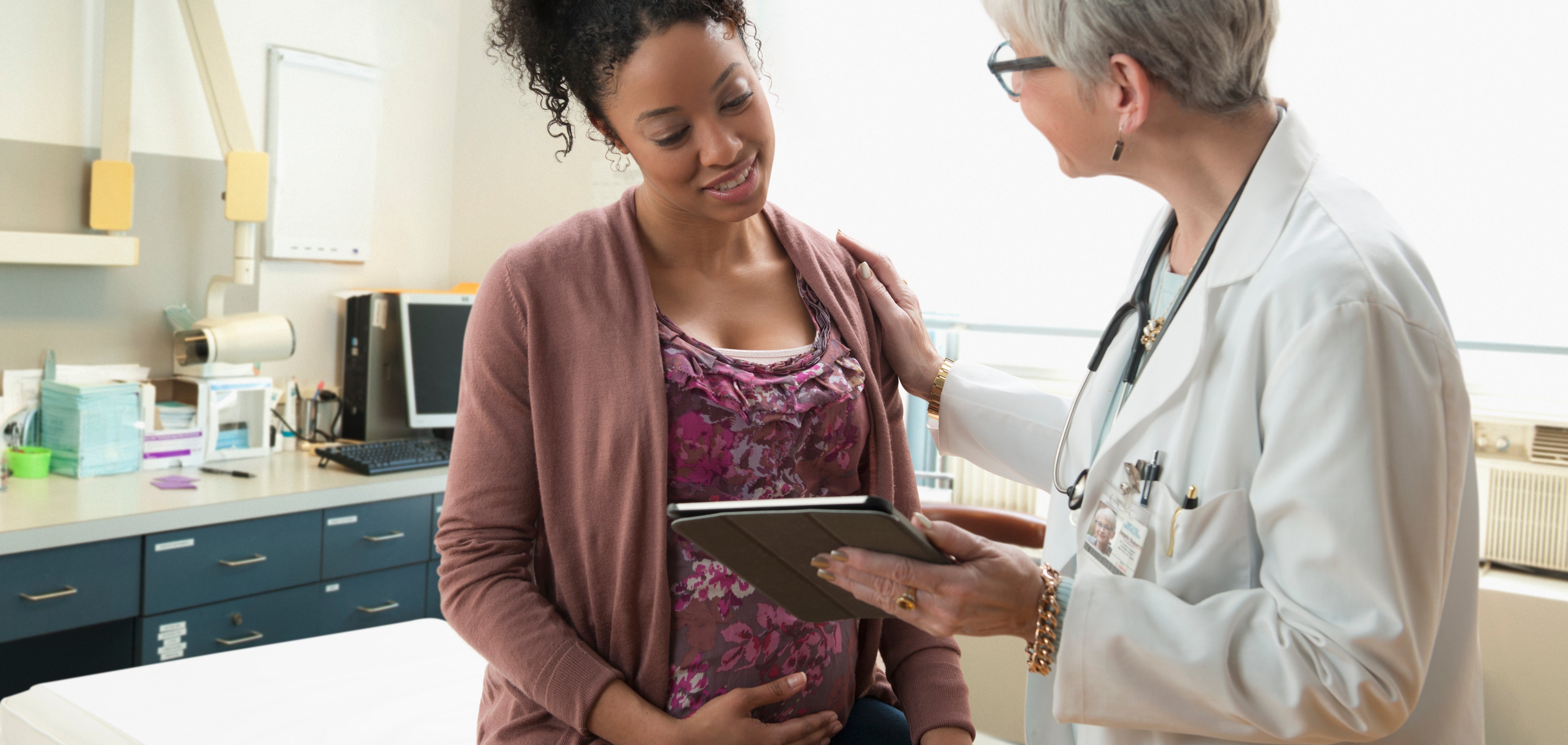 Lege i en laboratorium som diskuterer med en gravid pasient som sitter på et eksamensbord, holder et nettbrett