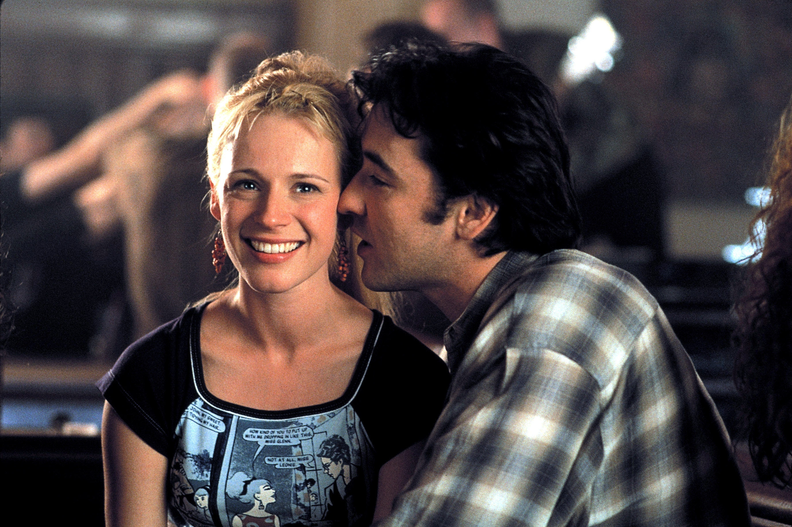 A woman smiles at the camera while a man in a plaid shirt leans in to whisper