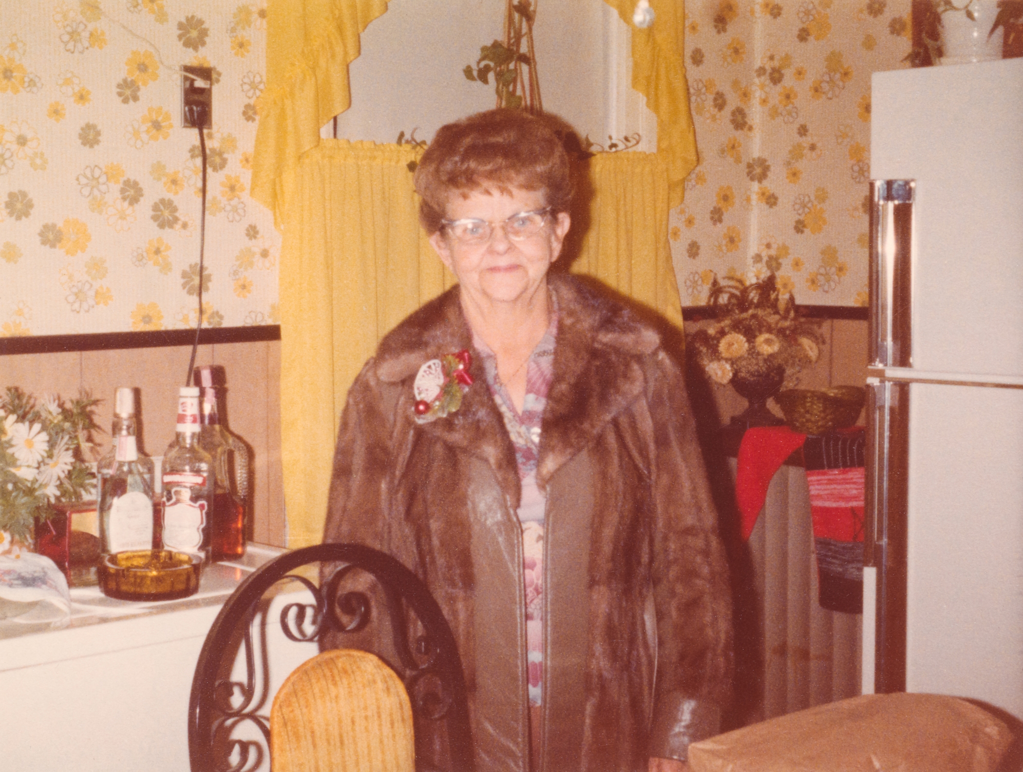 Eldre kvinne i en pels står på et vintage kjøkken med blomsterbakgrunns- og brennevinflasker på benkeplaten