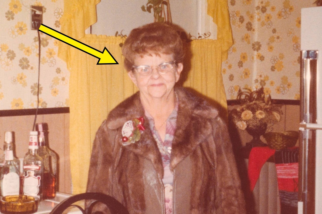 An older woman with glasses and a fur coat stands in a vintage kitchen with floral wallpaper and bottles on a counter