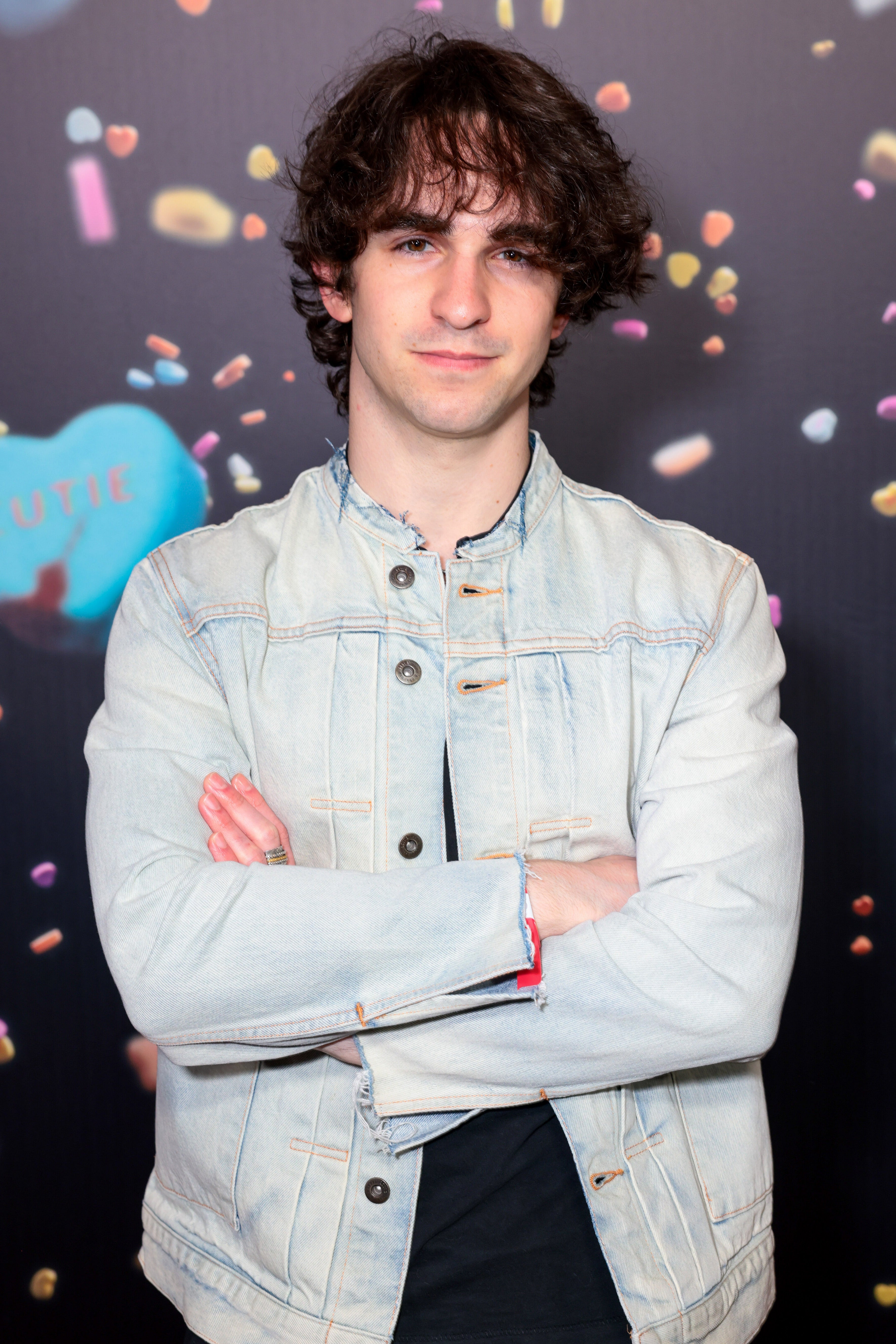 Person posing with arms crossed, wearing a light denim jacket. Background features abstract shapes and colors