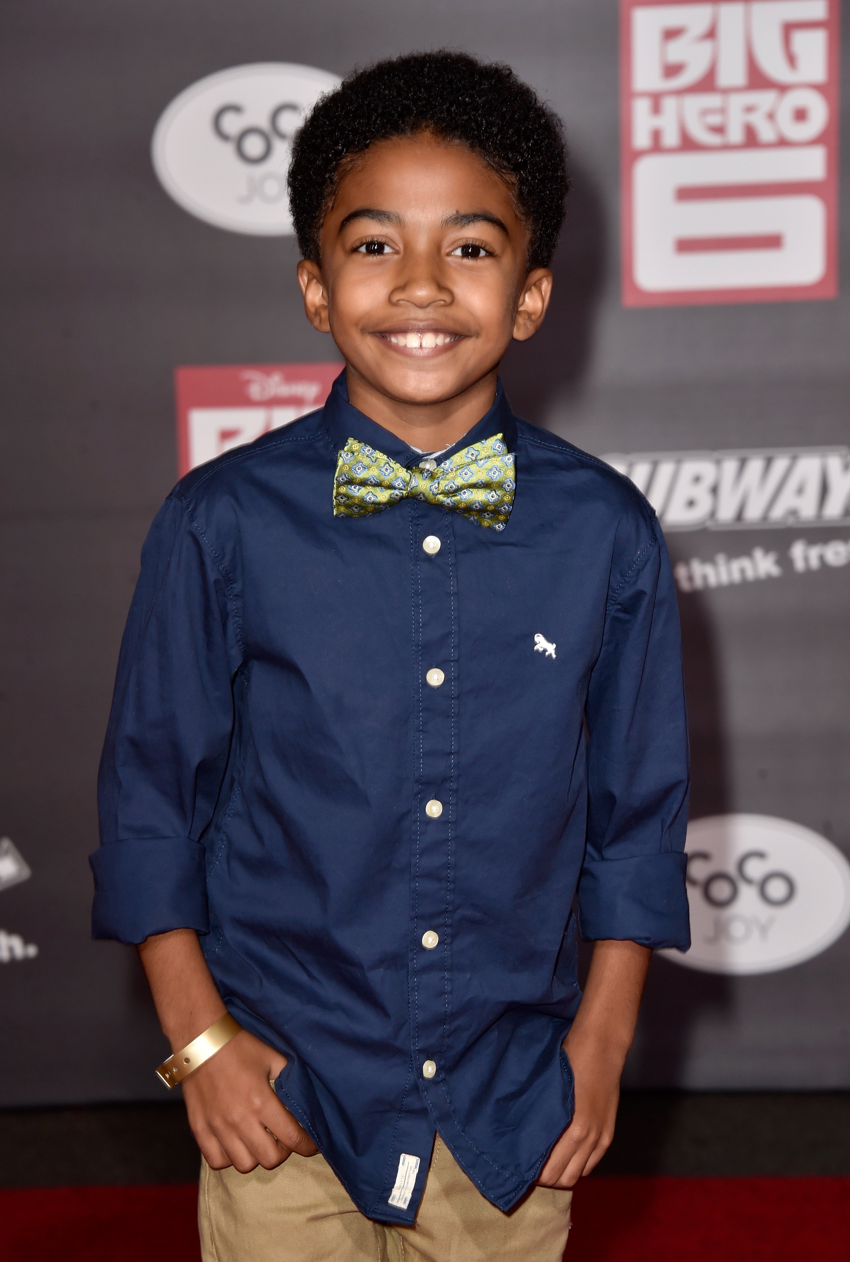 Young person smiling on the red carpet, wearing a navy shirt with rolled-up sleeves, a patterned bow tie, and khaki pants