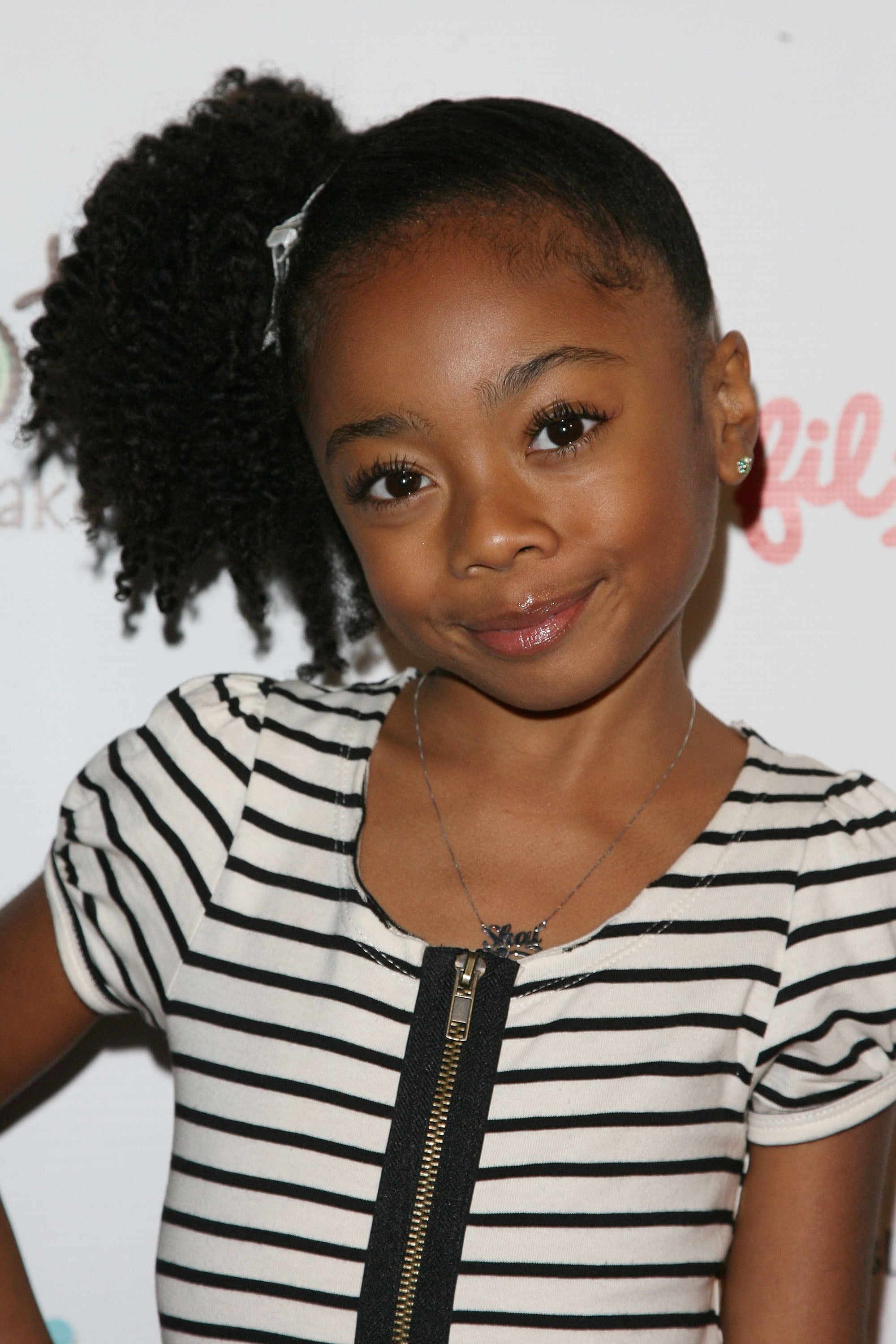 Young girl poses in a striped dress with a zipper, styled hair, and a necklace, smiling at a media event