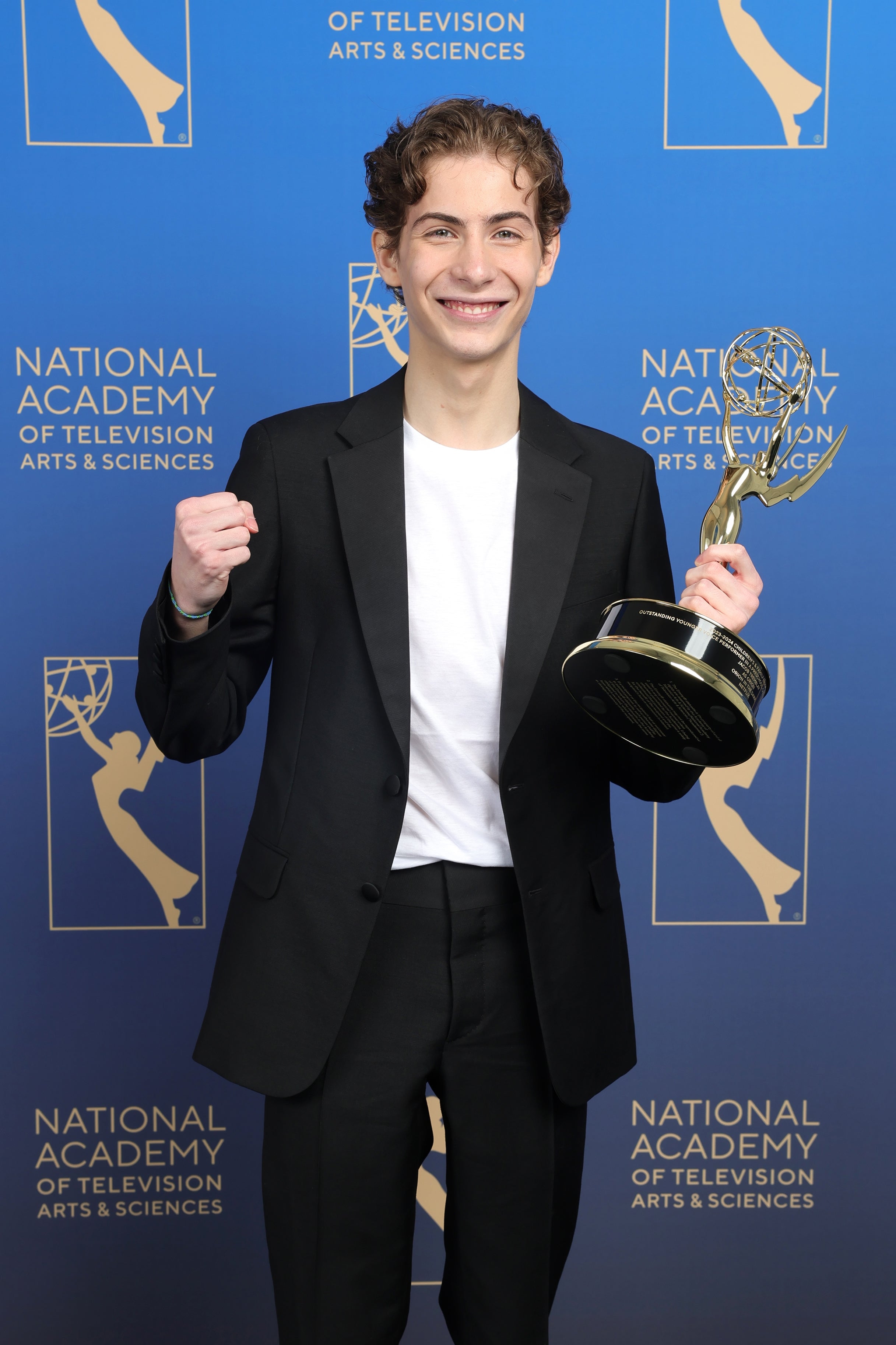 Person in a suit holding an Emmy Award, standing before a National Academy of Television Arts &amp;amp; Sciences backdrop, smiling with a fist raised
