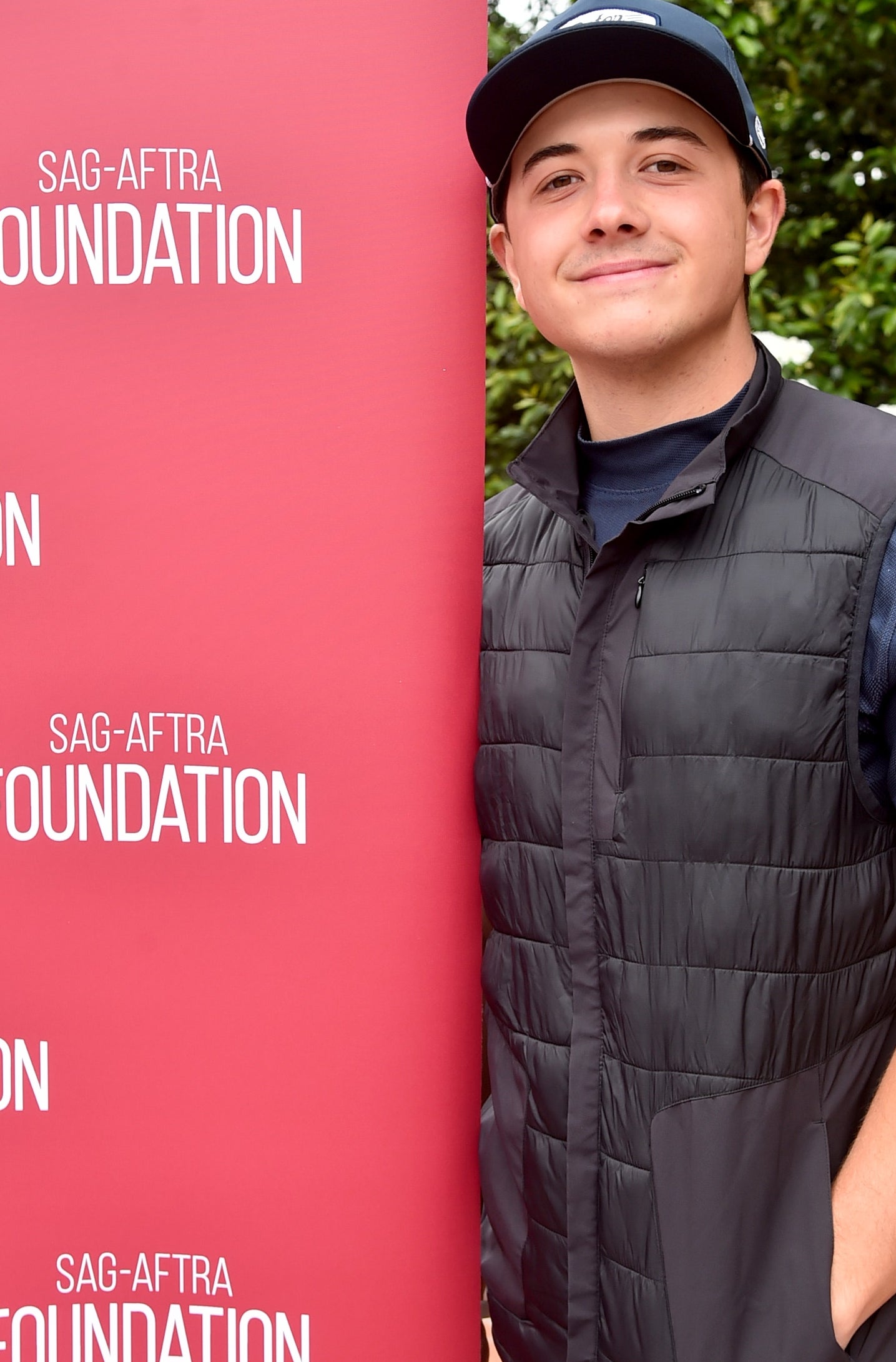 Person standing next to a SAG-AFTRA Foundation sign, wearing a casual cap and vest, smiling gently at the camera
